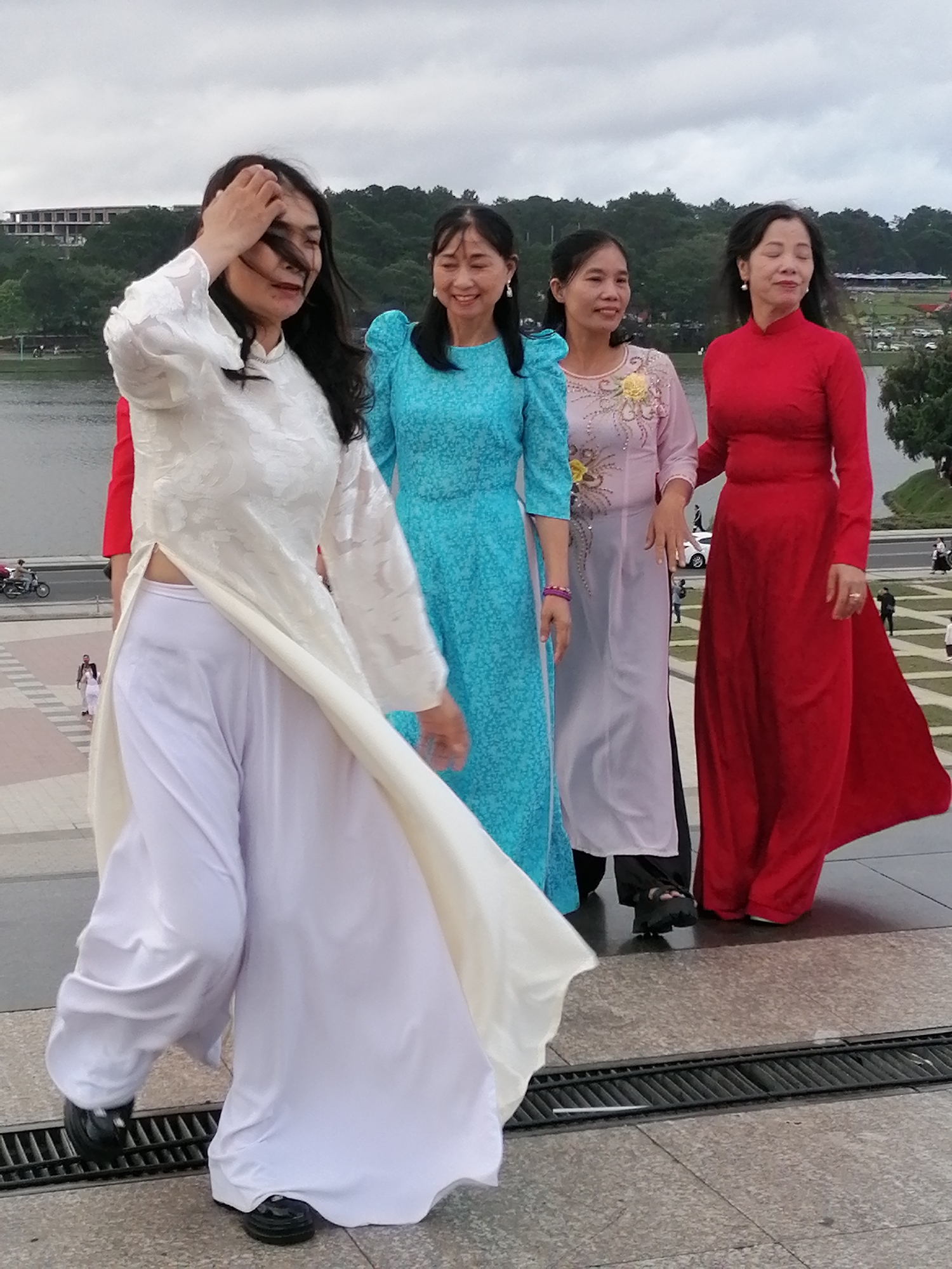 Four women stand in front of a body of water on an overcast day, with a hillside covered in trees and some buildings in the background. The women are dressed in traditional Vietnamese attire, known as Áo dài. The woman on the far left wears a white Áo dài over white pants, her hand is raised to her hair. The second woman from the left is wearing a bright blue Áo dài. The third woman from the left is wearing a light pink Áo dài with floral embroidery. The woman on the far right is wearing a vibrant red Áo dài. The setting appears to be a public outdoor area in Da Lat, Vietnam, possibly near Xuan Huong Lake, a well-known landmark in the city. The ground in the foreground is paved with stone, and a metal grate runs across the surface.