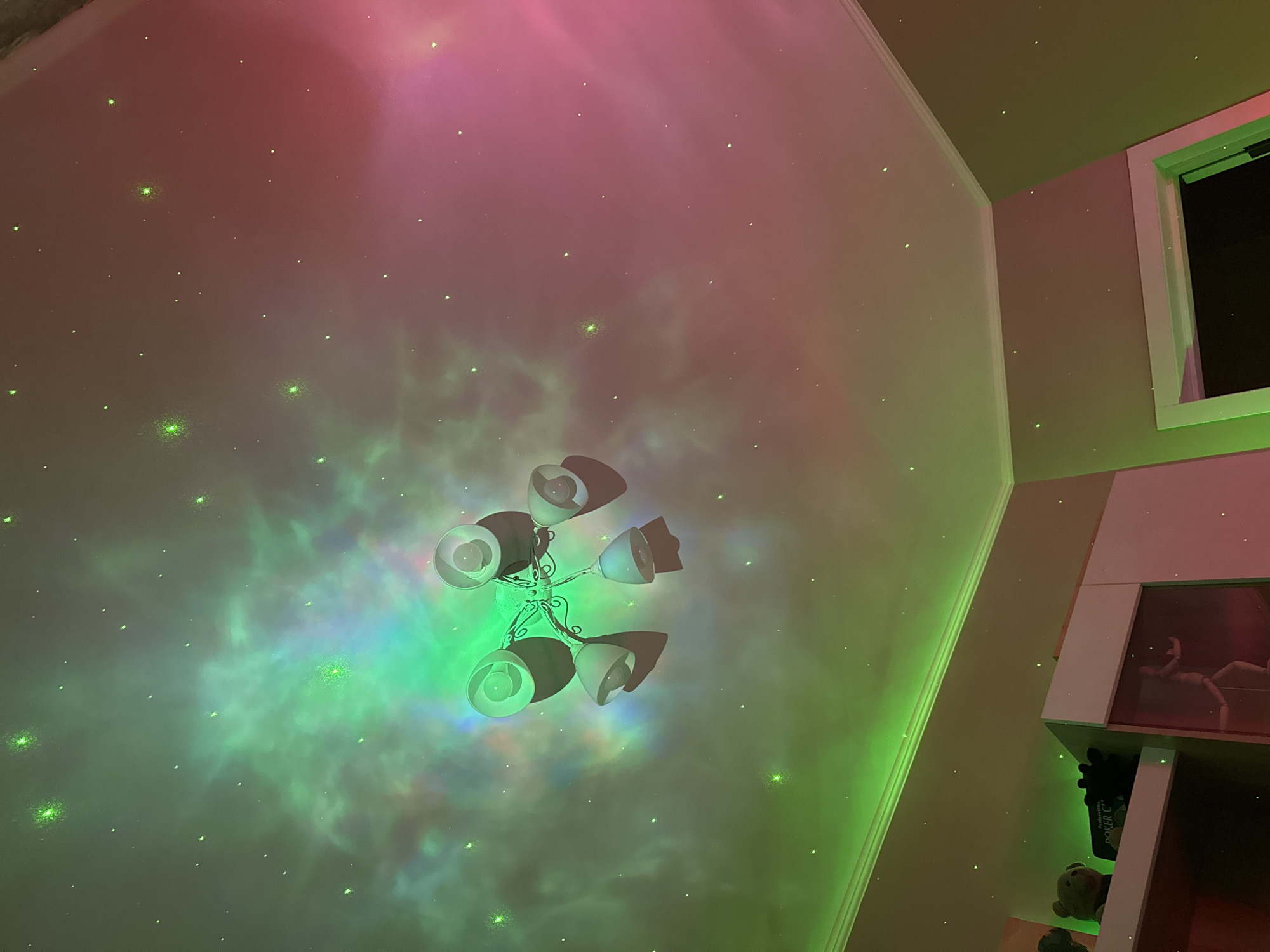 A low-angle interior shot shows a ceiling painted with a nebula pattern of pink and green hues, dotted with small green stars. A light fixture with five lamps is centrally located. The wall on the right side of the frame is a light neutral color with a white-framed window in the upper-right corner. Below the window is a white shelving unit with visible items, including a wooden mannequin, a green box that says "Poker C," and a teddy bear. The lighting casts a neon glow on the room.