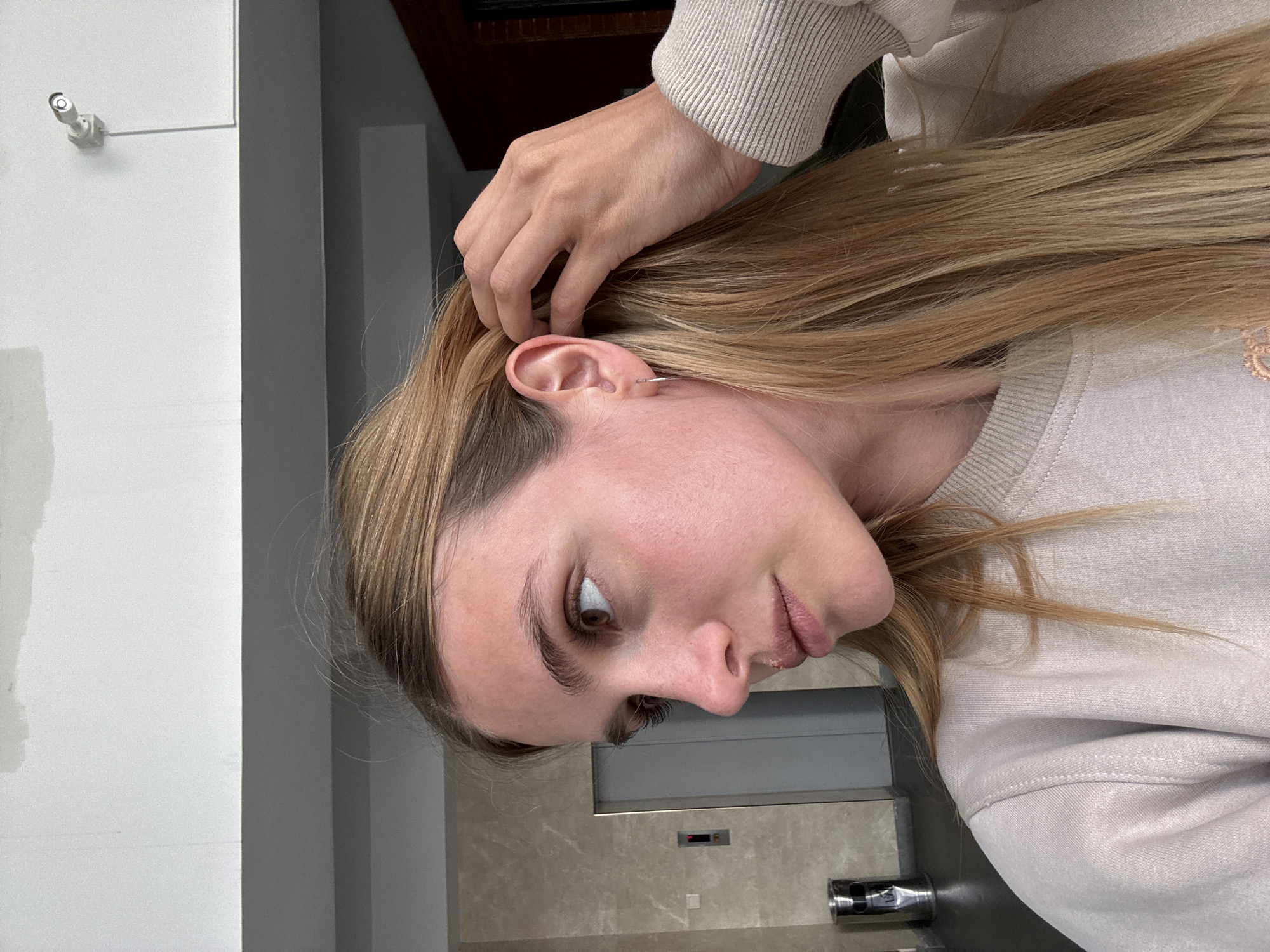 A woman with light brown hair, wearing a light-colored sweatshirt and small hoop earrings, is looking at the camera. Her right hand is near her ear, touching her hair. In the background, there is a white wall with a mounted camera, a gray wall, and a marble-patterned wall in what appears to be a modern interior space. While the image itself doesn't provide specific location details, there is no information in the image that could confirm it is from the city of Tbilisi, Georgia.