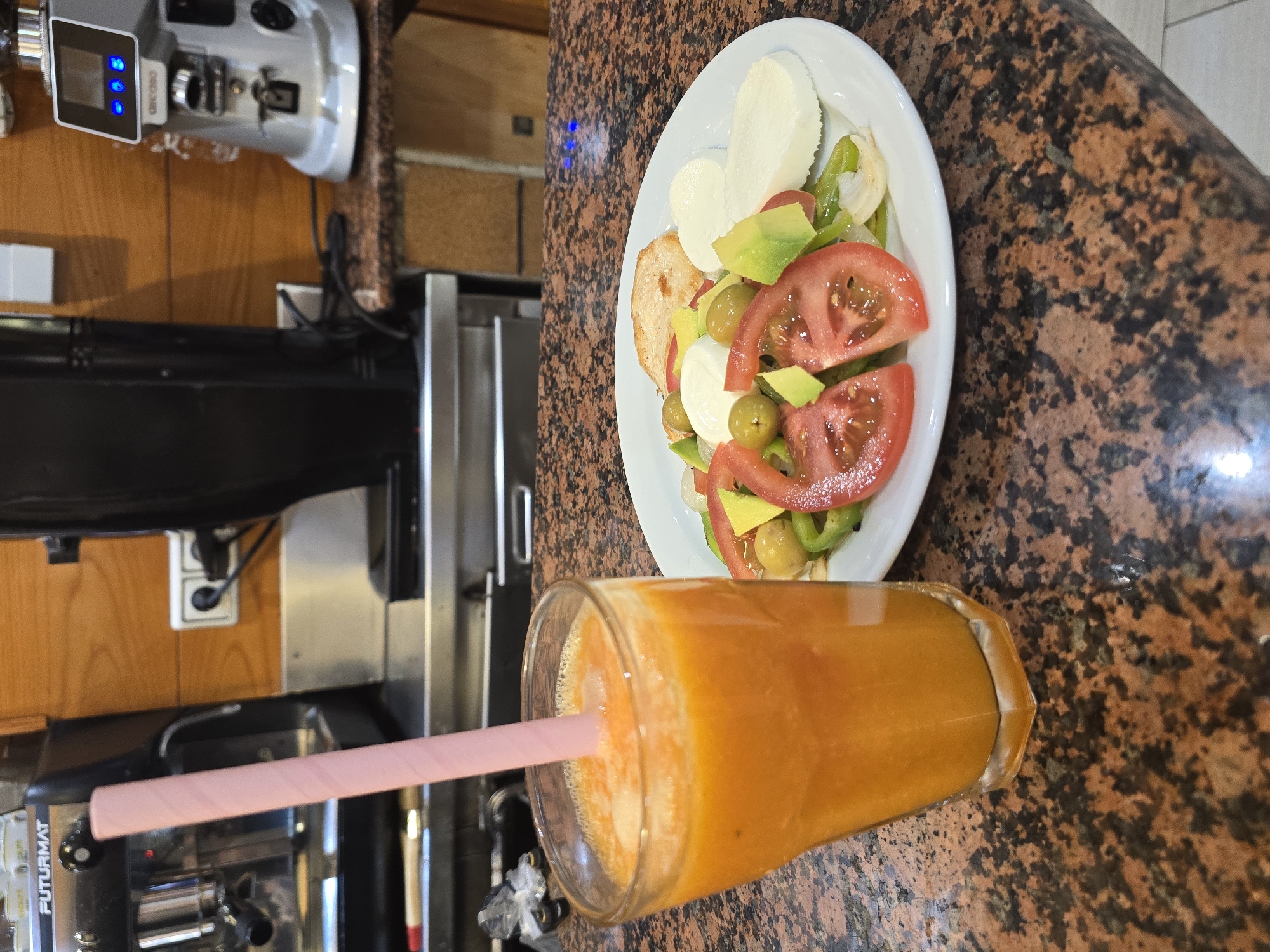 A close-up, eye-level shot displays a glass of orange juice with a pink straw on a speckled brown countertop. To the right, a white plate holds a salad with sliced tomatoes, green bell peppers, olives, avocado, and white cheese, along with a piece of toasted bread and chicken. In the background, a commercial espresso machine is visible on the left, and a silver coffee grinder is on the right. The environment suggests a cafe or restaurant setting in Barcelona, Spain.