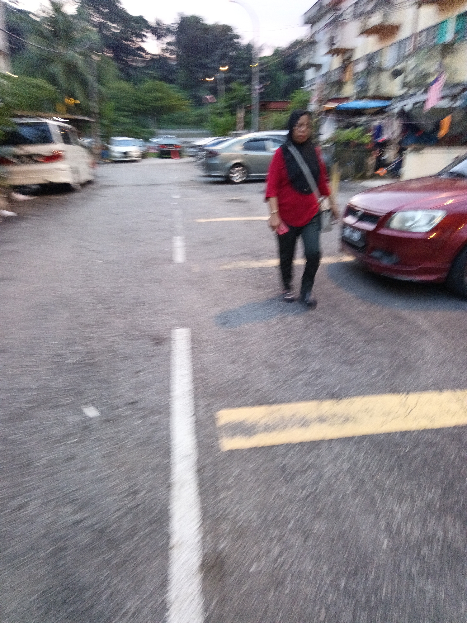 In Kuala Lumpur, Malaysia, on a paved road with white and yellow lane markings, a woman wearing a red top and black hijab walks alongside a red car. The woman has a bag slung over her shoulder and is holding a pink object. In the background, several cars are parked or driving, including a white minivan and a silver sedan. Buildings with balconies and flags are visible in the distance. The lighting suggests it is either early morning or late evening.