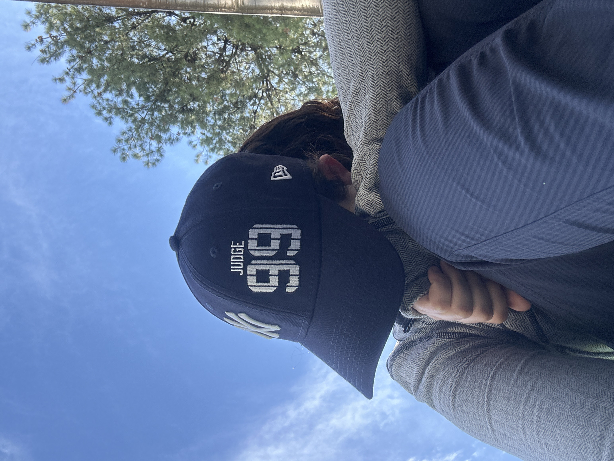 The image shows a person wearing a navy blue baseball cap with the New York Yankees logo and the name "JUDGE" and number "99" embroidered in white. The person is wearing a grey herringbone pattern jacket and a dark blue collared shirt. The sky is blue with scattered clouds. The setting is outdoor, with a tree partially visible in the upper left corner of the image. There is no information available to confirm the location is in Puebla, Mexico.