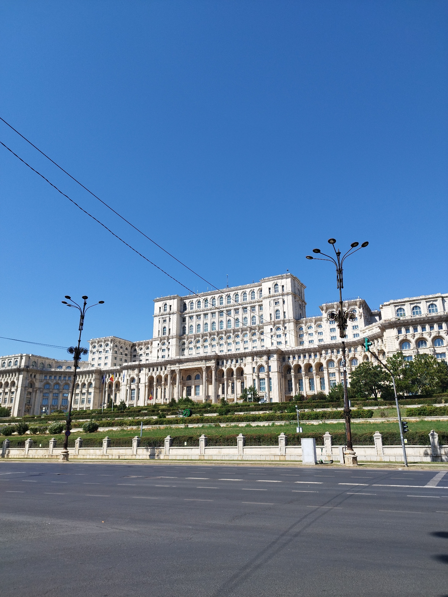 The image shows the Palace of the Parliament in Bucharest, Romania. The colossal neoclassical building, constructed from white stone and adorned with numerous arched windows and columns, dominates the frame. It is situated on a terraced hillside, with manicured greenery and decorative fencing in front. A wide asphalt road with white lane markings occupies the foreground, with streetlights and overhead power lines visible against a clear blue sky. The scene is devoid of people or vehicles, suggesting a quiet moment in a public space.