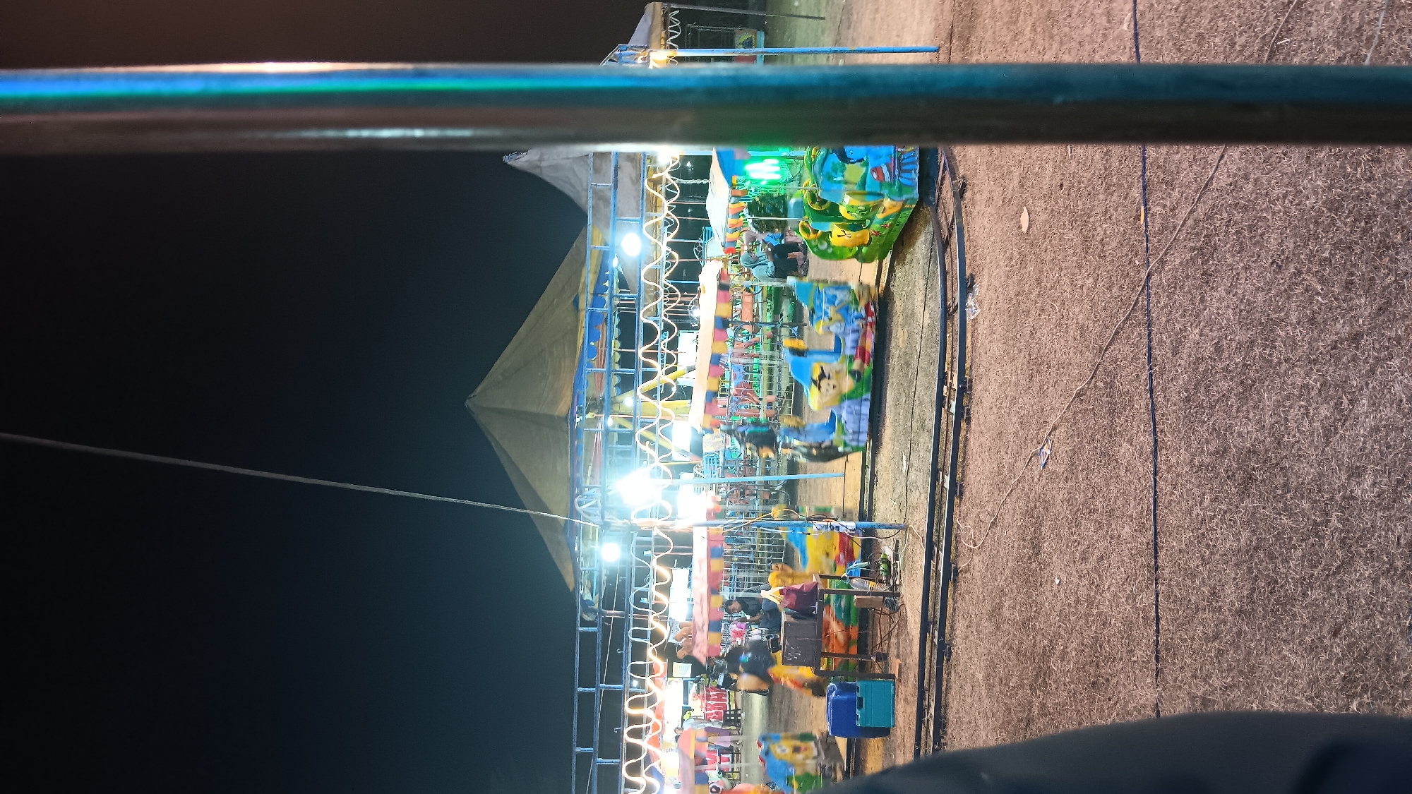 The image captures a nighttime scene at a small amusement park or fair in Getas, Indonesia. A miniature train ride is in motion, with colorful, cartoon-themed carriages on a track. Several people are visible, including a few seated on the train rides, and others standing or walking in the background. The amusement park is set up outdoors, with tents and a central structure that may be a carousel or another ride, illuminated by strings of lights. The ground appears to be covered in dry grass or soil, and power cables are laid out across the area. A thick metal pole, likely part of the amusement park infrastructure, is prominently in the foreground on the right side of the image, partially obscuring the scene. The overall atmosphere suggests a lively local event under the night sky.