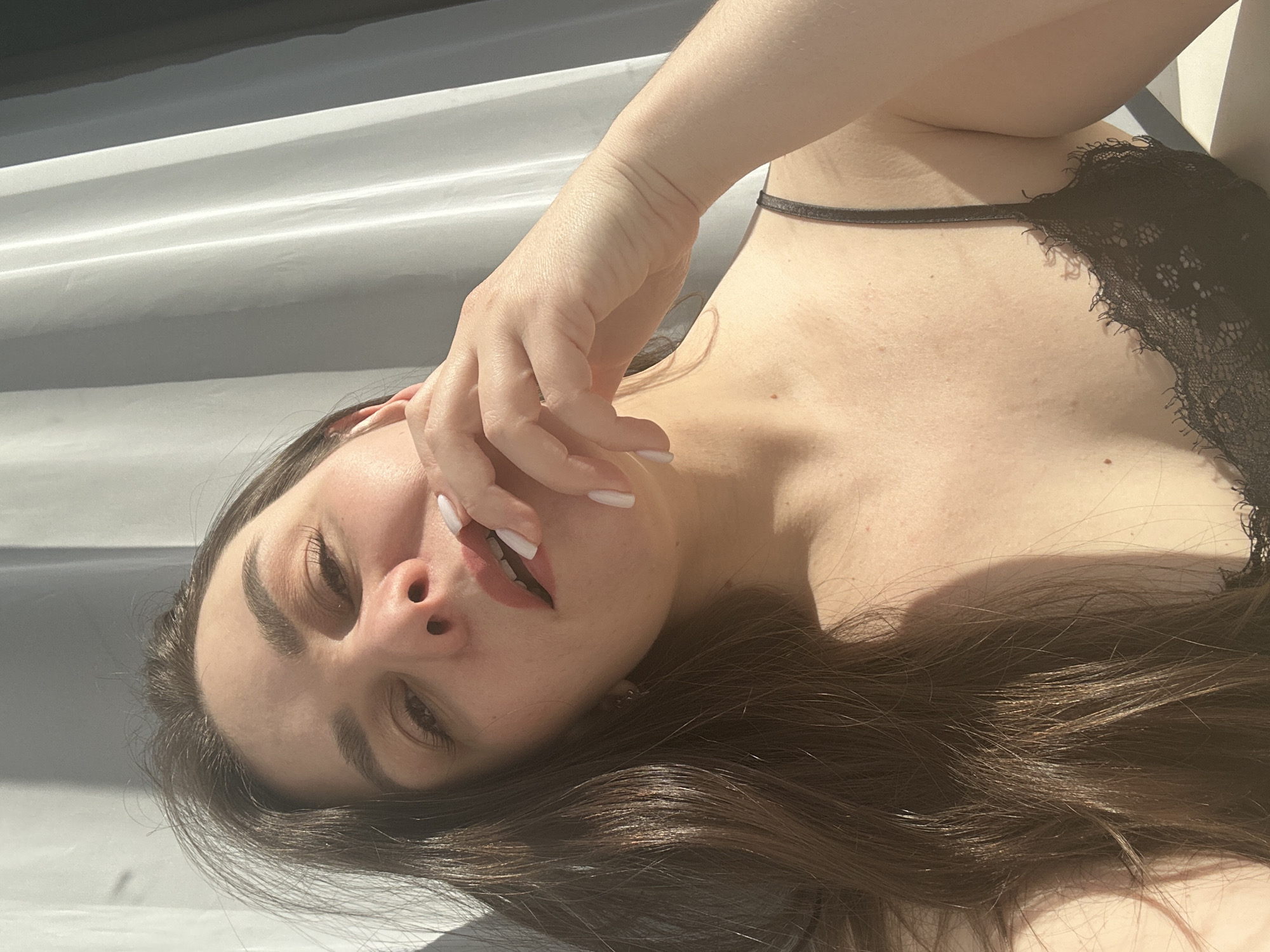 The image captures a close-up, diagonal view of a young woman bathed in bright, direct sunlight. Her head is positioned towards the lower left of the frame, with her body extending diagonally upwards to the right. She has long, dark brown hair, which spreads out around her head and partially covers her shoulder. Her facial features include well-defined eyebrows, partially closed eyes, and full lips, with her mouth slightly open, revealing a glimpse of her teeth. Her expression appears relaxed and introspective. Her right hand, featuring white-painted fingernails, is delicately placed near her chin, with fingers lightly brushing her skin.
She is wearing a black lace bralette or top, the delicate lace pattern visible on her upper torso. Her skin tone is fair, with a few faint freckles visible on her chest. The background consists of light-colored, horizontal, ridged surfaces, creating a subtle striped pattern, possibly a wall or paneling. The strong sunlight casts sharp shadows across her body and the background, highlighting the contours and textures. No text is visible in the media file.