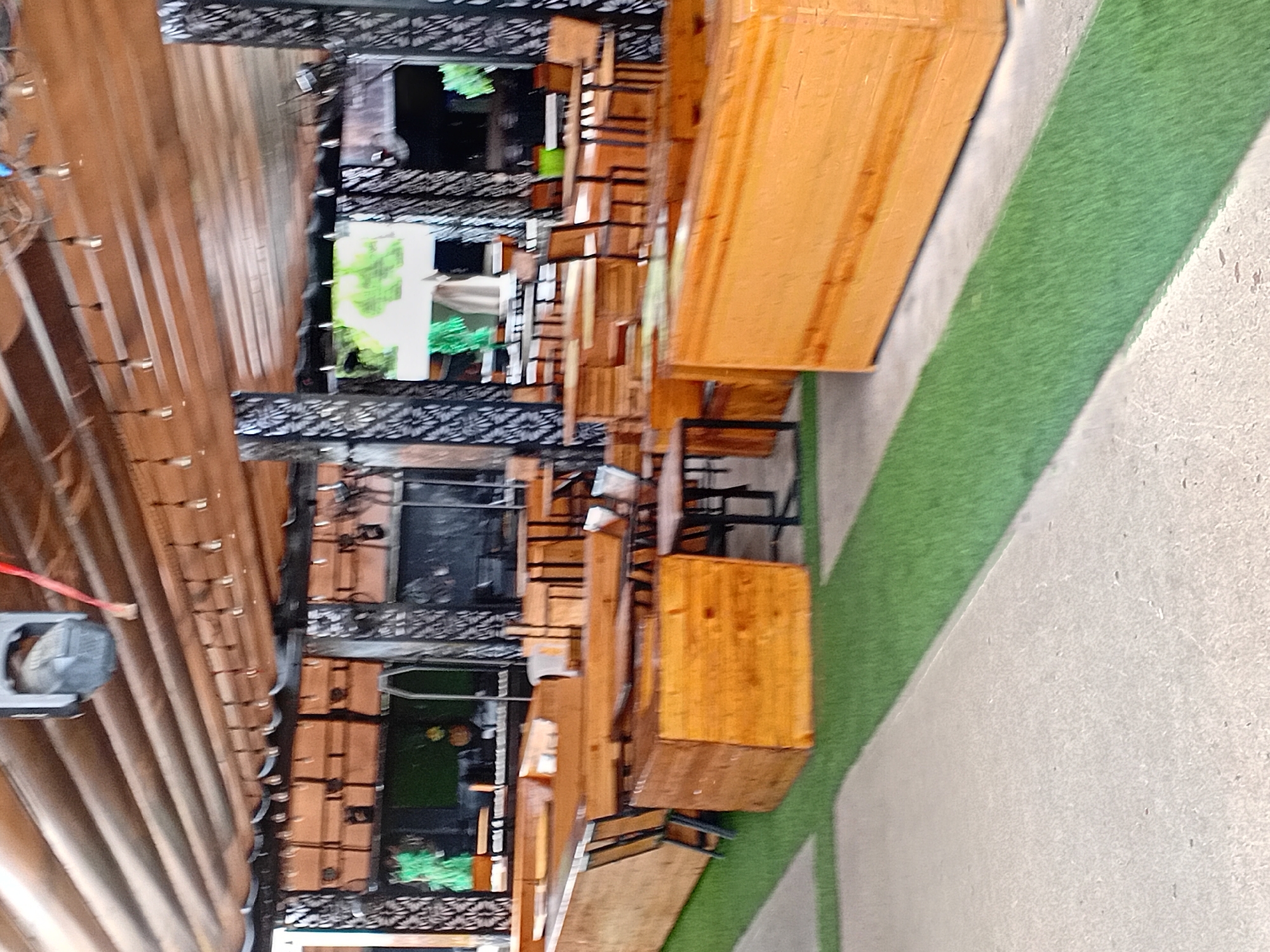 This is a medium shot, outdoor photograph of a restaurant or bar seating area. The seating is made of wooden benches and tables. There are also some chairs. The floor is covered with green turf, and there is a concrete walkway. The ceiling is made of wooden planks and there are lights hanging from it. The restaurant appears to be empty. The location is identified as a restaurant in the city of Howayi, Nigeria.