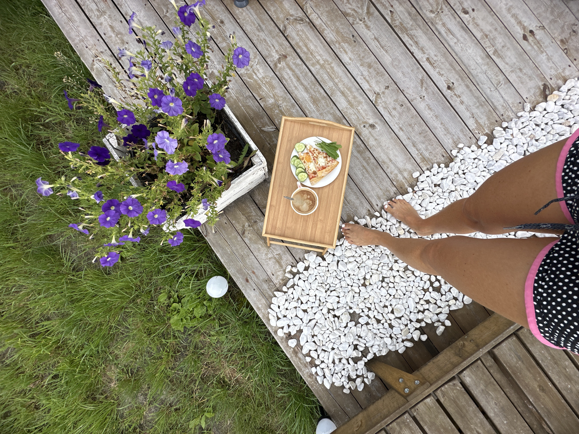 An aerial shot shows a woman's legs from the knees down, wearing black and pink polka dot shorts, standing on a weathered wooden deck. She is positioned to the right of a pile of smooth white stones. To her left, a wooden tray with a cup of coffee and a slice of pizza topped with cucumber slices and rocket leaves rests on the deck. In the background, a white planter box overflows with vibrant purple petunias. The deck extends towards a patch of lush green grass. Two white spherical garden lights are visible on the grass. The scene suggests an outdoor setting, possibly a backyard or patio.