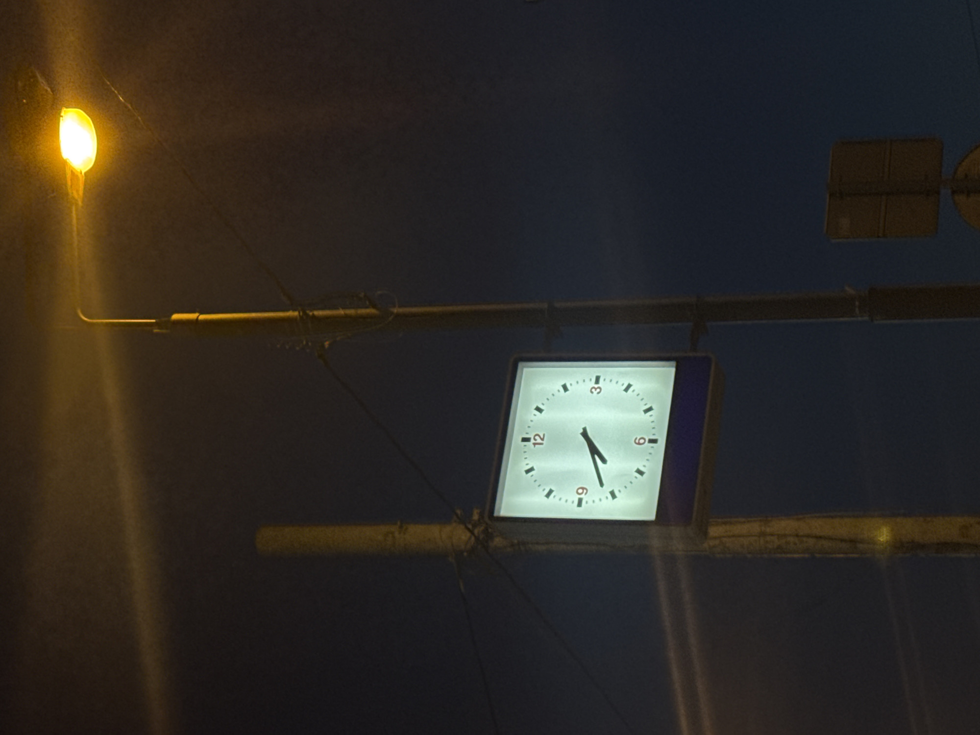 The image shows a clock mounted on a metal support, likely part of a street lamp or utility pole, at night. The clock face is illuminated, displaying a time of approximately 4:33 or 4:34. A street lamp is visible to the left of the clock. The environment appears to be an urban area in Moscow, Russia.