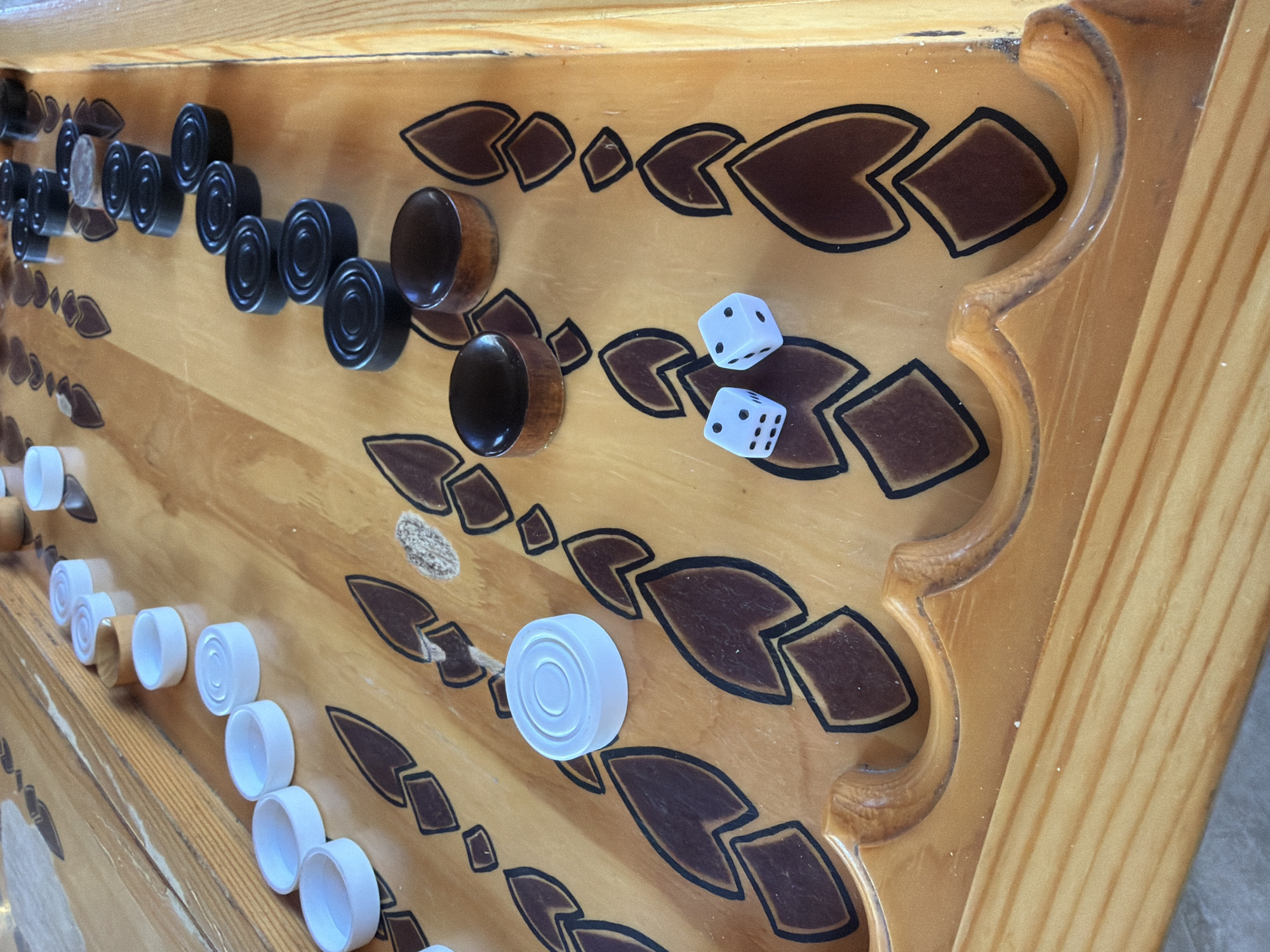 A high-angle, perspective shot shows a backgammon board. The board has a light brown wood finish with dark brown and black decorative markings. There are black checkers arranged in a row on one side of the board, and white checkers arranged in a row on the opposite side. Two white dice rest near the center of the board. The location is Таскино, Russia.