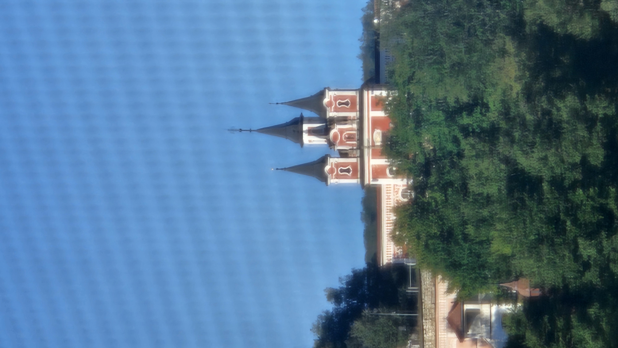 The image shows a red and white church with multiple spires against a bright blue sky. The church is situated on a hill surrounded by lush green trees. In the foreground, more trees obscure the lower portion of the structure. A stone wall with ornate detailing runs along the front of the building. This is the Orthodox Cathedral of Saint John the Baptist, located in Prešov, Slovakia.