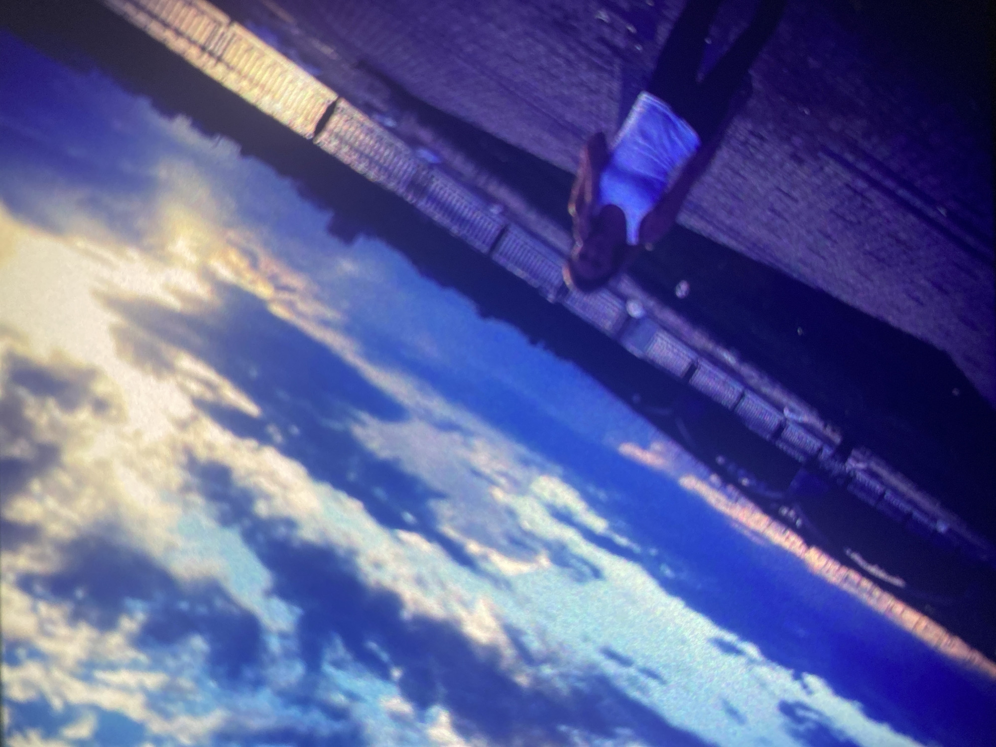 The image displays a dramatically tilted perspective, with a vibrant sky filling the lower-left and a large structure featuring a person dominating the upper-right. The sky is awash with colors characteristic of either sunrise or sunset, transitioning from bright yellow and orange near a hidden horizon to deep blue, with scattered white and grey clouds.
A prominent, dark, and seemingly metallic structure, possibly a bridge or an elevated walkway, runs diagonally across the upper portion of the frame. It appears to have a grated or textured surface. On this structure, a person is visible, positioned in the upper-right. They are wearing a light-colored top, possibly white, which exhibits a striking, almost neon-blue glow, and dark trousers or leggings. Their head is visible, looking generally downwards or towards the camera's perspective, though facial details are obscured by shadow and distance.
The scene suggests an elevated location, possibly a bridge or high platform, offering views over the landscape or water beneath a colorful sky in Saint Petersburg, Russia. The notable blue luminescence on the person's shirt could be caused by artificial lighting, UV light, or strong reflections from the blue sky or water. No text is visible in the image. The overall impression is one of dramatic lighting and an unusual vantage point.