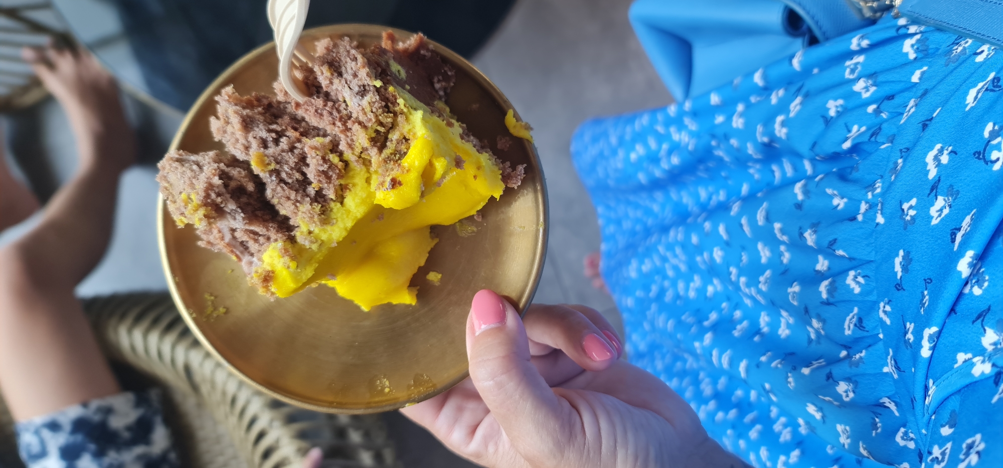 The scene shows a partially eaten slice of brown cake with bright yellow frosting on a small, round, golden plate being held by a person with pink nail polish. The person is wearing a blue dress with white floral patterns and is partially visible from the chest down. Beside them is a bare foot, and in the blurred background, there is a leg wearing a blue and white patterned garment. The background also contains a wicker-style piece of furniture. The image was likely taken from an elevated position. There is no information provided in the image to suggest it was taken in Đà Nẵng, Vietnam.