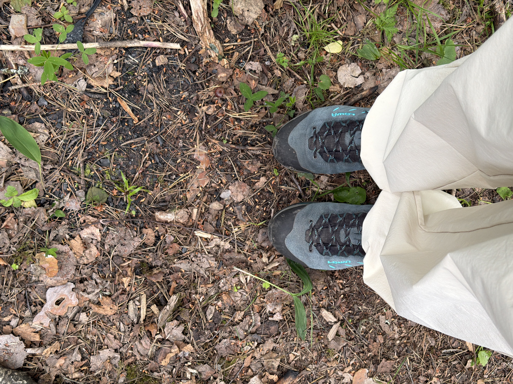 The image shows a top-down view of a person's legs and feet standing on a forest floor. The person is wearing light-colored pants and dark gray hiking boots with teal laces and accents. The ground is covered with fallen leaves, pine needles, twigs, and some patches of green vegetation. The scene appears to be in a natural outdoor setting, likely a forest or wooded area. The location is specified as Kuznechnoye, Russia.