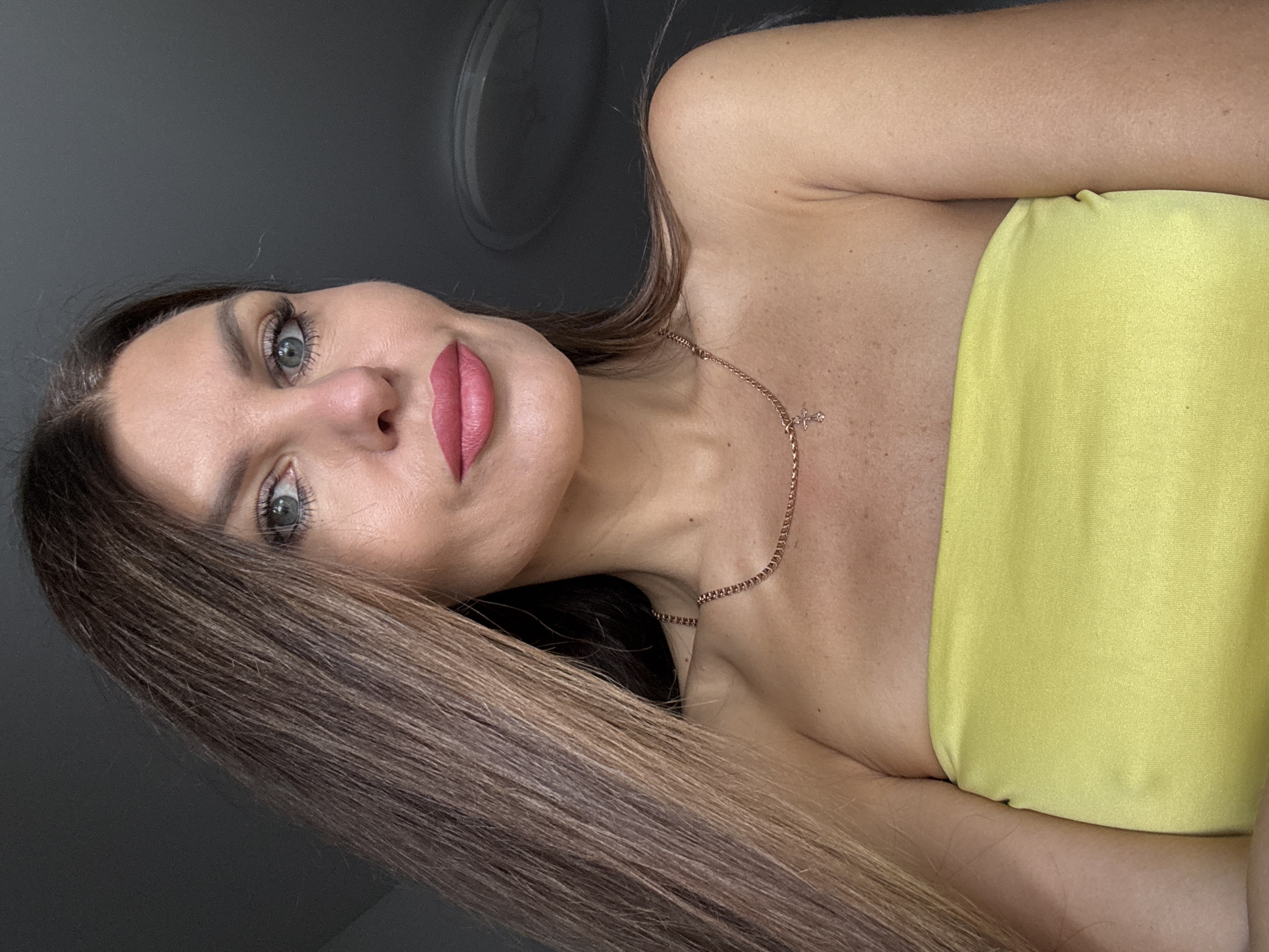 The image shows a close-up of a woman with long, brown hair. She is wearing a strapless yellow top and a delicate gold necklace with a cross pendant. The woman has blue eyes and pink lipstick. The background is dark and indistinct, suggesting an indoor setting. The image was taken in Chaikovsky, Russia.