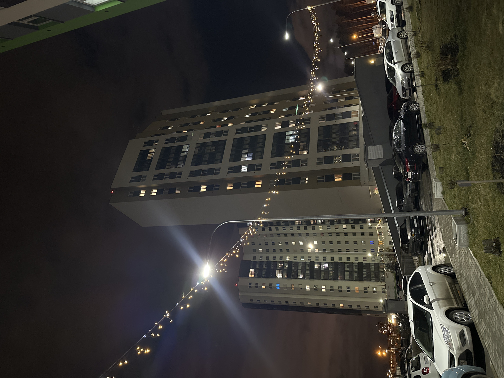 This media file presents a night scene in a residential area of Yekaterinburg, Russia. The primary subjects are two tall, modern high-rise apartment buildings, their facades marked by numerous rectangular windows, many of which are lit, suggesting habitation. The architecture features contrasting light-colored panels and darker window sections, with one building exhibiting a more grid-like window pattern and the other having larger, more distributed window units.
In the foreground and extending along the right side, a parking area with several parked cars is visible. These vehicles include white, black, and other dark-colored cars, with one white sedan partially identifiable as a Ford Focus in a cropped view. Patches of grass and paved walkways border the parking spaces.
The scene is brightly illuminated by multiple streetlights, which create noticeable lens flares. A string of decorative, warm-toned fairy lights spans across the upper portion of the image, adding a festive or aesthetic element to the otherwise functional lighting. The sky is dark with visible cloud cover, indicating it is nighttime. There are no people visible, and no specific activities are explicitly taking place, portraying a quiet, everyday evening in an urban residential complex. A car's license plate, partially visible in one of the provided crops, shows the regional code "96," corresponding to the Sverdlovsk Oblast where Yekaterinburg is located.