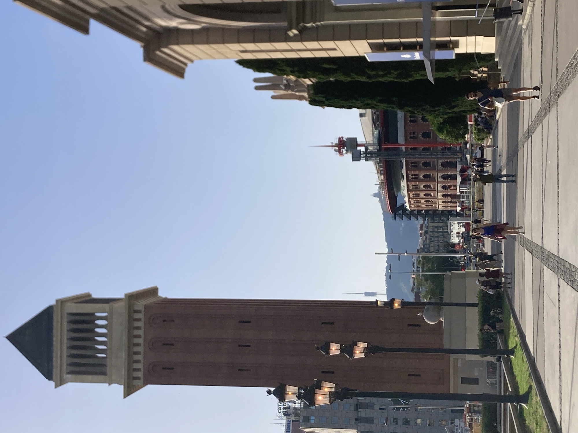 The image depicts a sunlit urban scene in Barcelona, Spain, likely at the Plaça d'Espanya. In the foreground, a wide, paved walkway with patterned sections extends into the distance. Several people are walking along this path. On the left, a tall, reddish-brown brick tower with a distinctive crenellated top stands prominently. Several ornate lampposts are positioned along the walkway near the tower.
To the right of the walkway, a section of a building with classical architectural elements, including arches and columns, is visible. Further into the midground, a large, circular, red-roofed building, resembling a bullring or arena, is prominent. A tall communication tower with a red antenna is situated behind it. Beyond these structures, the cityscape of Barcelona stretches out, with numerous buildings and a distant hill or mountain range. The sky is clear and blue, indicating a bright, sunny day. The overall impression is one of a lively public space, possibly a tourist attraction or major public square within the city.