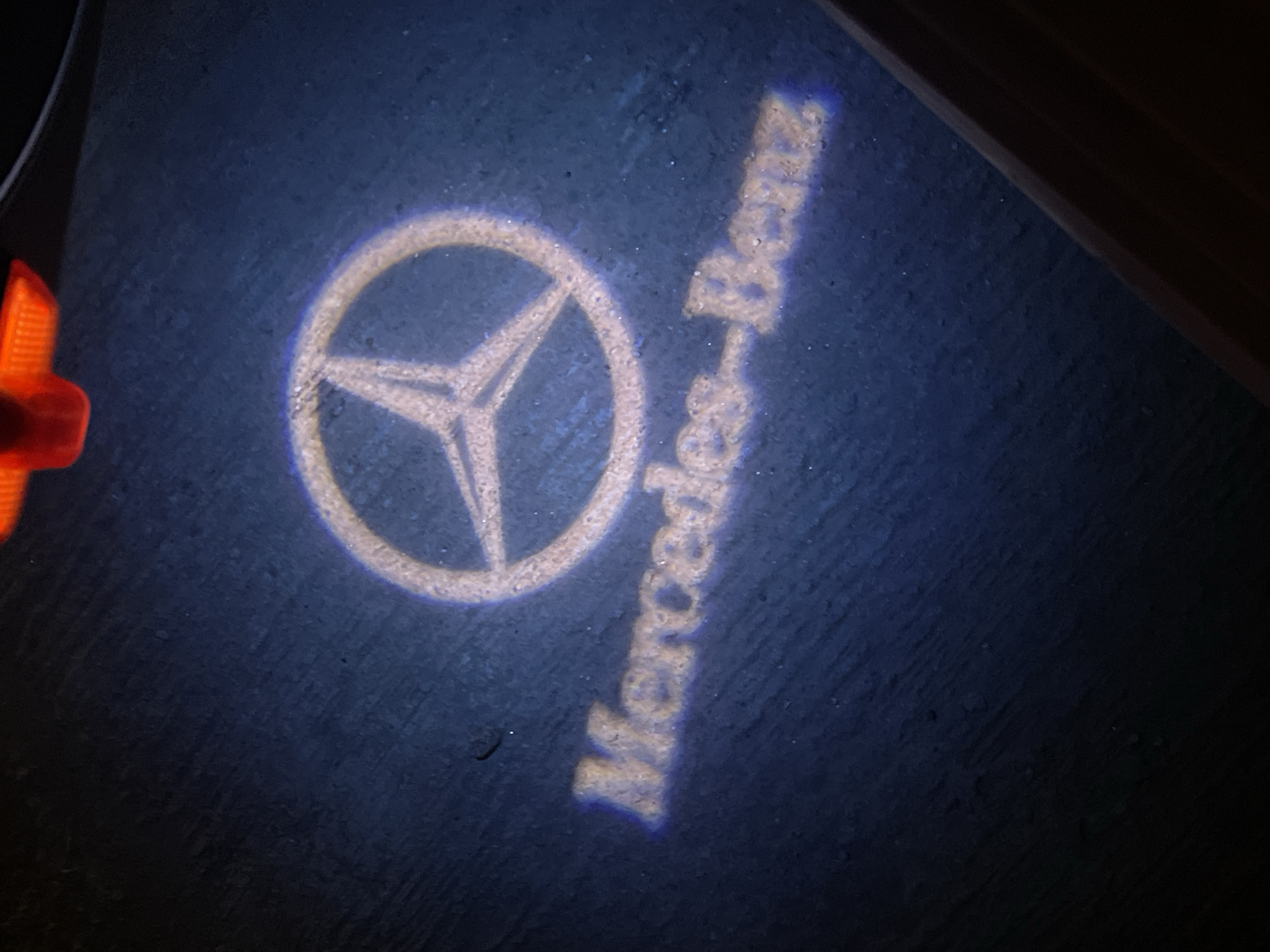 The image shows a Mercedes-Benz logo and the words "Mercedes-Benz" projected onto a dark surface, likely the ground. A portion of an orange object is visible in the upper left corner. There is insufficient information to determine the location within the city of Abuja, Nigeria.