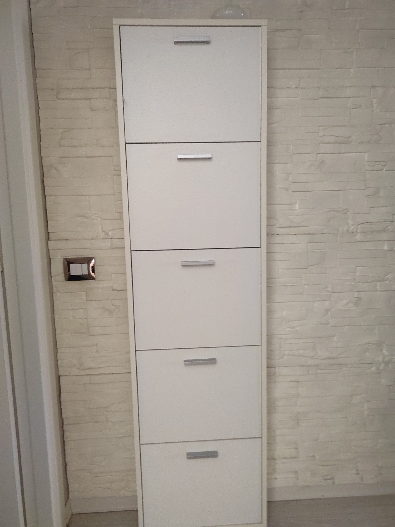 The image shows a tall, narrow, white shoe cabinet with five drawers, each with a silver handle. The cabinet is positioned against a light-colored wall with a textured, brick-like pattern. To the left of the cabinet, a portion of a white door frame is visible. Below the second drawer from the top, there is a light switch with a silver frame and white toggles, set into the brick-patterned wall. The floor is a neutral color. The image appears to be taken indoors, likely in a hallway or entryway. There are no people or animals present. The setting is consistent with an apartment in Porto Recanati, Italy.