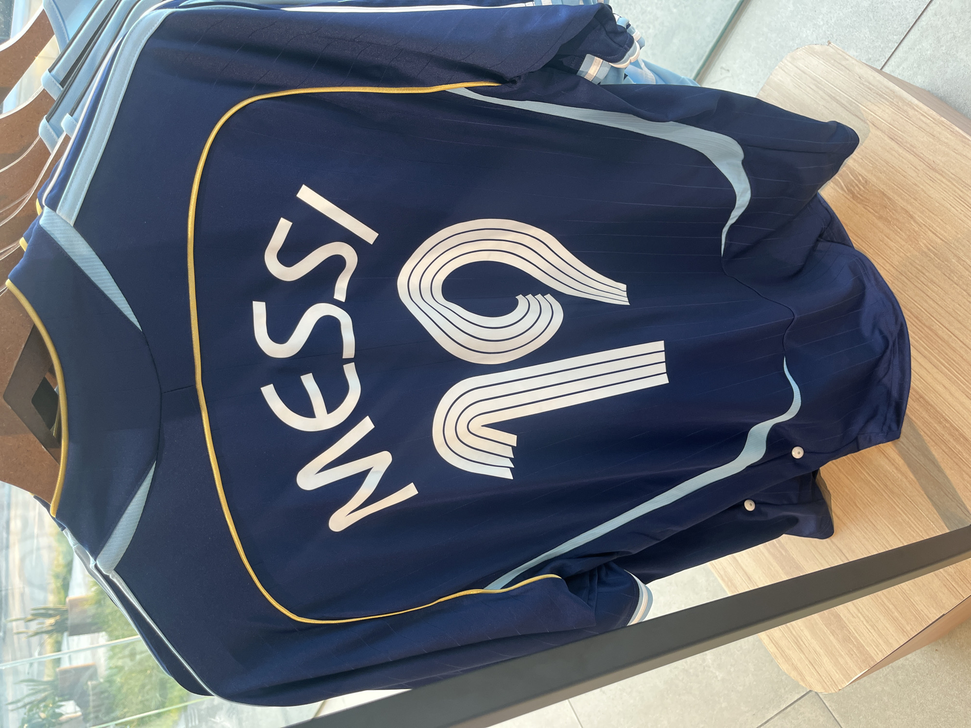 A close-up shot shows the back of a navy blue soccer jersey with the name "MESSI" and the number "10" printed in white. The number 10 is stylized with multiple parallel lines. The jersey features light blue and gold trim. The jersey is hanging on a rack. The location is inside a store.