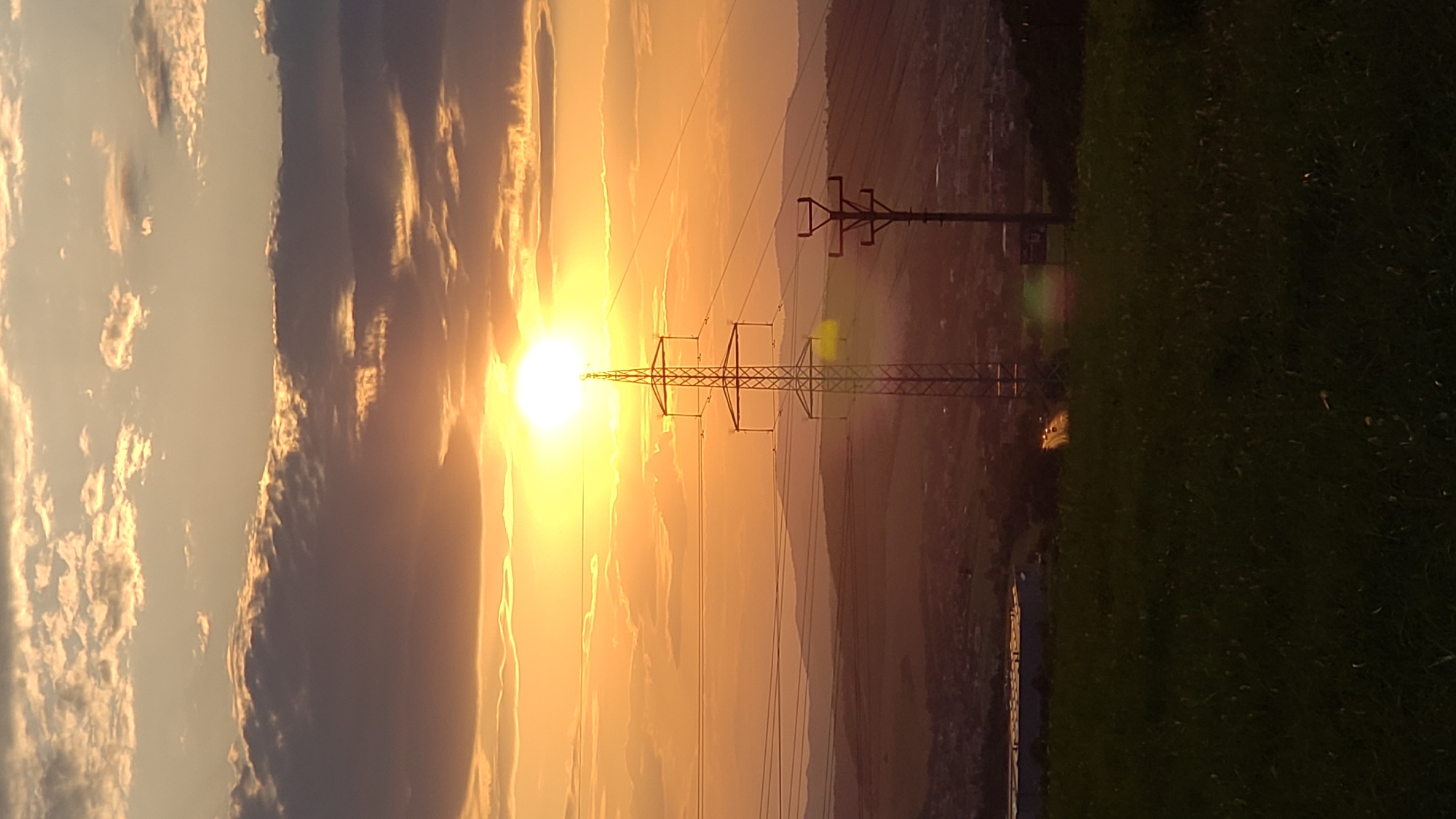 A vertical, high-angle shot captures a sunset over the city of Prešov, Slovakia. The sun, a bright orb, is positioned slightly left of center, surrounded by a halo of yellow light and partially obscured by wispy clouds tinged with orange. In the foreground, dark green grass dominates the bottom of the frame.
Dominating the mid-ground are two tall electricity pylons. The pylon on the left is larger and more prominent, constructed of lattice metal. To its right, a smaller, single-pole pylon stands. Power lines stretch horizontally across the frame, connecting these pylons and extending into the distance.
Beyond the pylons, the landscape slopes downwards, revealing the city of Prešov. Buildings are visible, though somewhat indistinct due to the distance and the fading light. A road with a few cars can be seen, their headlights creating streaks of light. In the far distance, rolling hills form a layered silhouette against the orange sky. The overall impression is one of a vast, natural landscape juxtaposed with human infrastructure, all bathed in the warm glow of a setting sun.