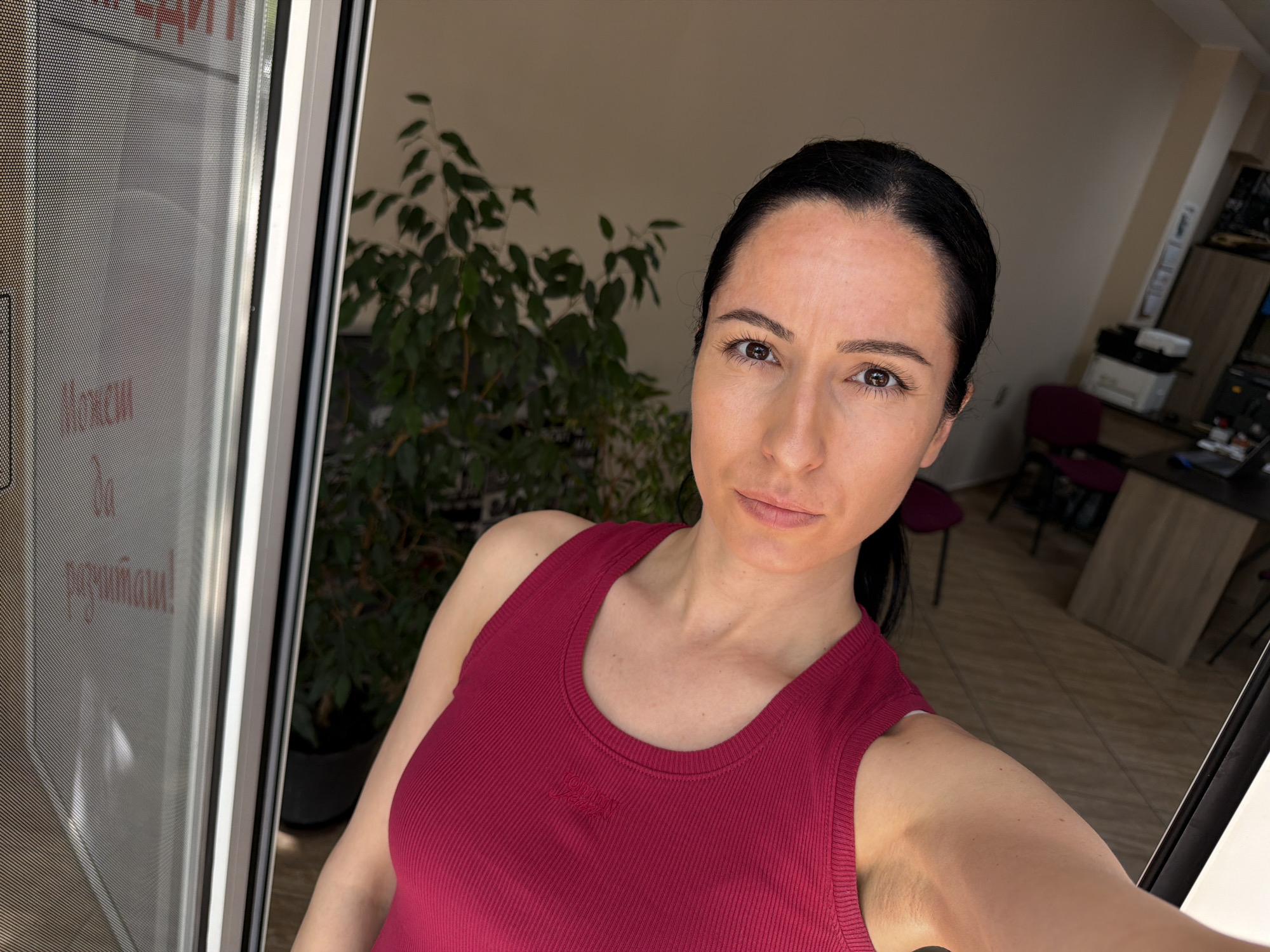 A woman is captured in a selfie, looking directly at the camera with a neutral, composed expression. She has dark hair pulled back, and her dark brown eyes are prominent. She is wearing a dark pink or magenta ribbed tank top.
The setting appears to be an indoor environment, likely a home or office in Pleven, Bulgaria. Behind her and to the left, a large, lush green houseplant, possibly a Ficus, adds natural greenery to the background. To her right, an adjacent room is visible, featuring what looks like an office setup with a light-colored desk, a printer/scanner, and a dark red office chair. Shelving units are also discernable on the far wall.
The woman is positioned near a glass door or large window panel, which is covered with a fine mesh screen. Visible on this screen, in Cyrillic script, is partial text that appears mirrored and potentially upside down. The decipherable portion reads "Макси да разчиташ!", which translates to "Maxi to count on!" or "You can count on Maxi!", suggesting a possible business name or advertisement relevant to the Bulgarian location. The overall lighting indicates it is daytime.