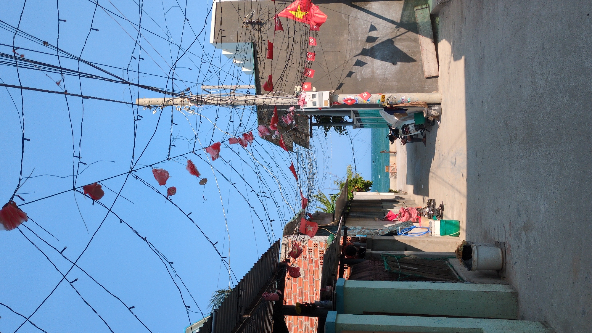 An alleyway in Phan Rang – Tháp Chàm, Vietnam, is depicted in this shot. Overhead, a dense network of electrical wires and strings adorned with small red flags spans the alley's width. A utility pole stands in the center of the frame, supporting the wire web.
The alley's right side features a concrete pathway, while the left side shows a mixture of residential structures constructed from brick, corrugated metal, and painted concrete. One or more individuals may be obscured by the structures on the left.
In the alley's distance, the sea is visible, suggesting proximity to the coast. A parked motorbike is also observed in the distance.
The sky above is clear and blue.