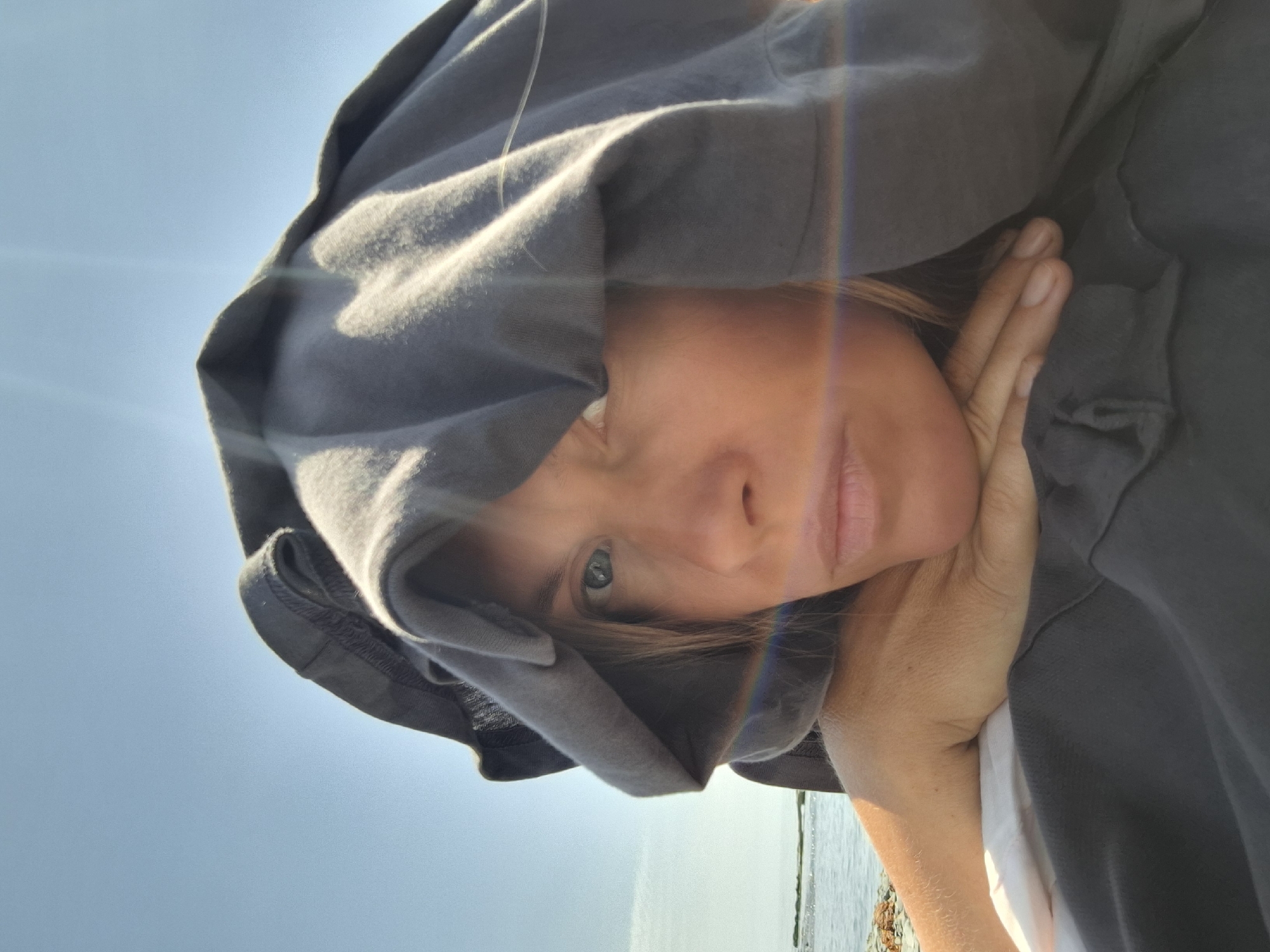 An extreme close-up selfie shows a woman's face partially obscured by a dark gray hooded sweatshirt. She is resting her head on her arm, with her hand supporting her chin. Her eyes are open and looking towards the camera. The background consists of a bright blue sky and a body of water, with hints of land or rocks in the distance. A subtle rainbow-like lens flare is visible across her face. The image appears to have been taken outdoors in natural light.