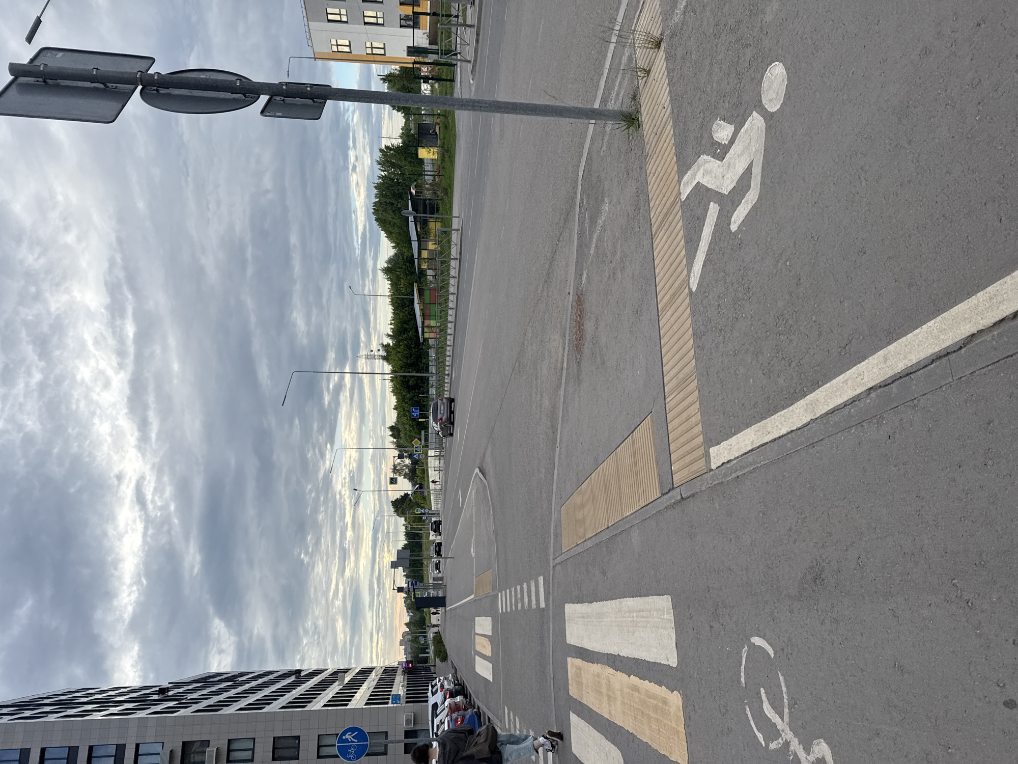 The image depicts a street scene in Perm, Russia. In the foreground, a pedestrian crossing is marked with white and yellow stripes. To the left of the crossing, a bicycle lane is indicated by painted symbols on the asphalt. A woman walks away from the camera on the sidewalk, carrying a backpack. In the background, a multi-story apartment building stands on the left, and a road with moving vehicles is visible. Traffic lights and street lamps are also present. The sky is overcast with clouds.