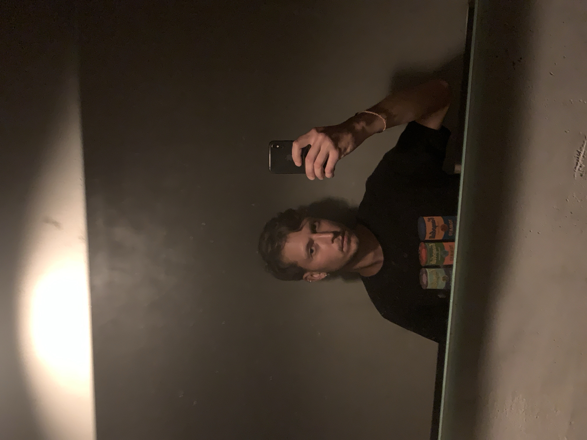 A man is taking a mirror selfie indoors. He is holding a black phone. He is wearing a black t-shirt and a bracelet. There are cans stacked on a shelf next to the mirror. The location is not specified and thus cannot be verified.