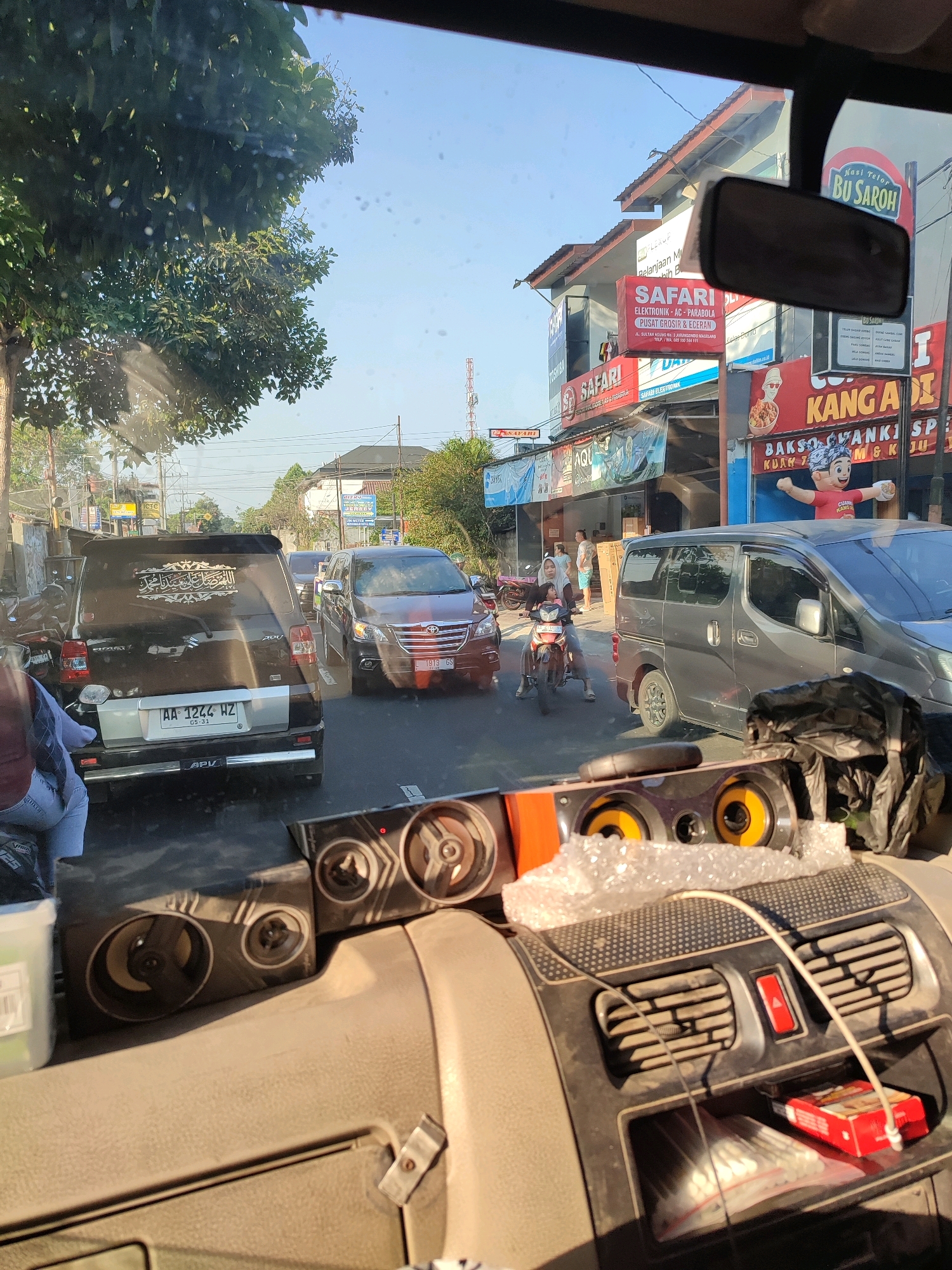 The image captures a street scene in Magelang, Indonesia, viewed from inside a vehicle. The foreground features part of a car's dashboard, with speakers, air vents, and a red cigarette pack visible. The view outside the windshield reveals a busy street with various vehicles, including a black SUV with Arabic calligraphy on its rear window, a dark red car, and a gray van. A woman and a child are seen riding a motorcycle. Buildings line the street, adorned with signs for businesses such as "Safari Elektronik," "Nasi Telor Bu Saroh," and "Kanga Adi." Trees and utility poles are also present. The sky is clear and blue, indicating daylight.