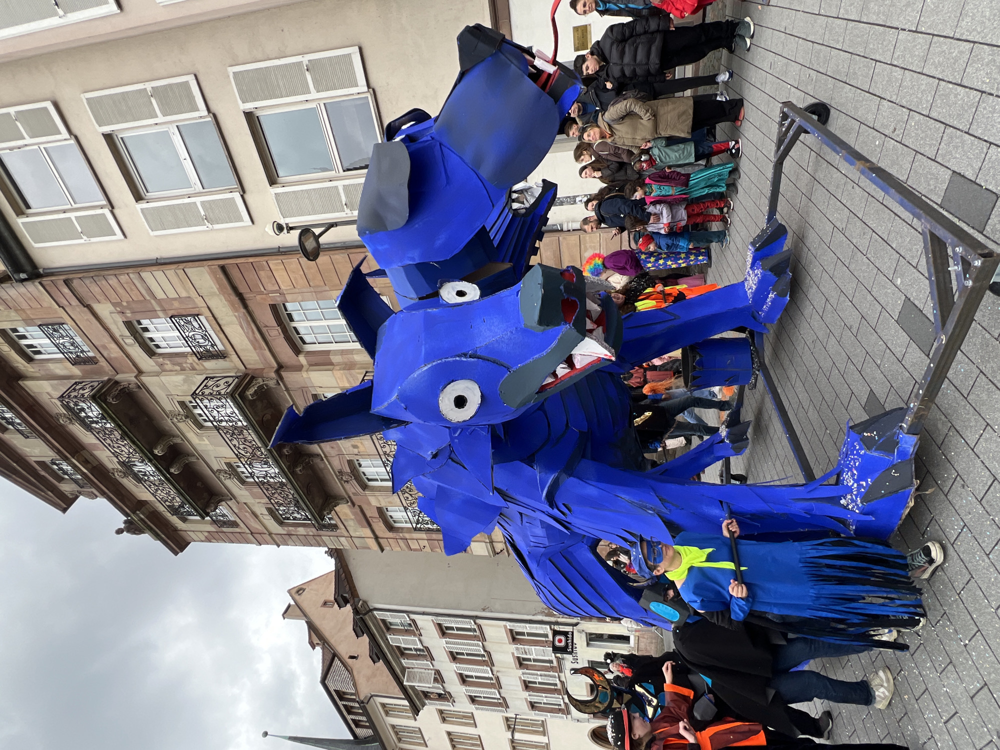 A sideways, wide-angle shot depicts a vibrant street scene in Strasbourg, France. The foreground showcases a large, cobalt blue, two-headed dragon-like figure constructed from cardboard or similar material. It is positioned on a wheeled frame pushed by a person in a blue costume. The dragon has a menacing expression with prominent white, circular eyes and pointed teeth.
A diverse crowd of onlookers lines the street, ranging from children in colorful costumes to adults in everyday winter attire. The people are watching or interacting with the parade or event.
The street is paved with gray bricks. Buildings on either side feature a mix of architectural styles, including windows with white shutters and more ornate facades with wrought-iron balconies. A sign for a Sushi restaurant is partially visible. The sky is overcast and gray.