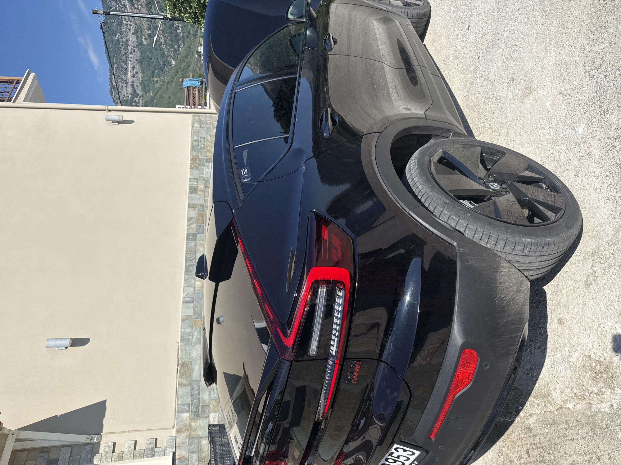 A black Volvo XC40 Recharge electric vehicle is parked outdoors on a paved surface, likely in a residential area. The car is positioned to show its rear and right side, with the hood of another vehicle visible in the background to the right. The car's taillights are illuminated, and the "Recharge" emblem is visible on the rear. The backdrop includes a building with light-colored walls and stone accents, and beyond that, a hilly landscape with trees under a clear blue sky. The scene suggests a sunny day. The license plate of the Volvo is partially visible and appears to be from Greece.