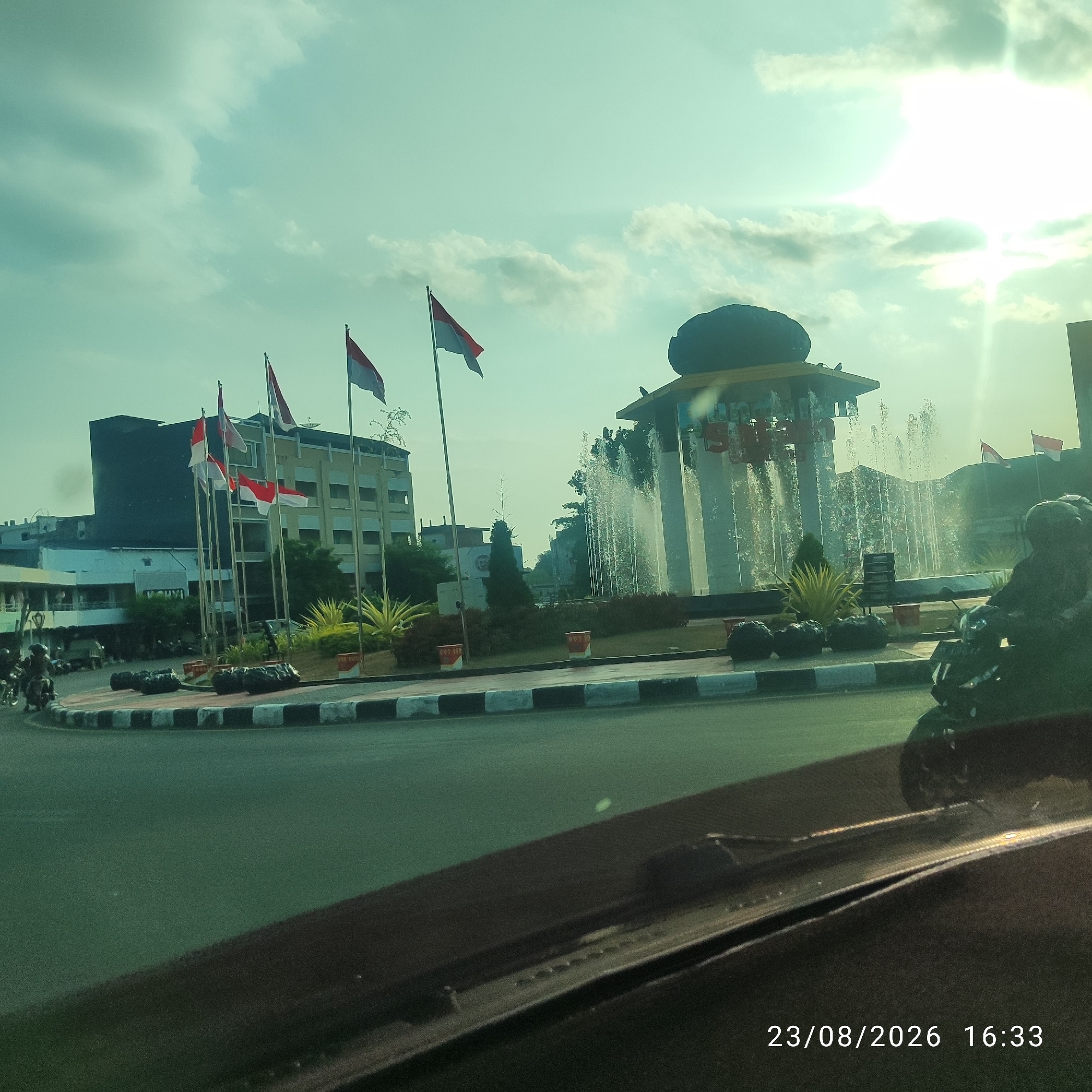 The image captures a street scene in Tanjung Pandan, Indonesia, dominated by a central roundabout featuring a fountain and several Indonesian flags. The fountain's water jets create a dynamic visual element against the backdrop of buildings and a bright sky with scattered clouds. Multiple flags, displaying the red and white colors of Indonesia, are prominently displayed on poles around the roundabout and on a building in the background. Several buildings, including a multi-story structure with dark-colored sections and a lighter, more decorative building, are visible. The foreground shows a portion of a vehicle's dashboard and windshield, suggesting the photograph was taken from inside a car. A motorcycle with a rider is visible in the right side of the frame, and other motorcycles are seen in the distance on the left. The date and time stamp "23/08/2026 16:33" is overlaid on the bottom right corner of the image. The overall environment suggests an urban setting with elements of national pride and public infrastructure.
