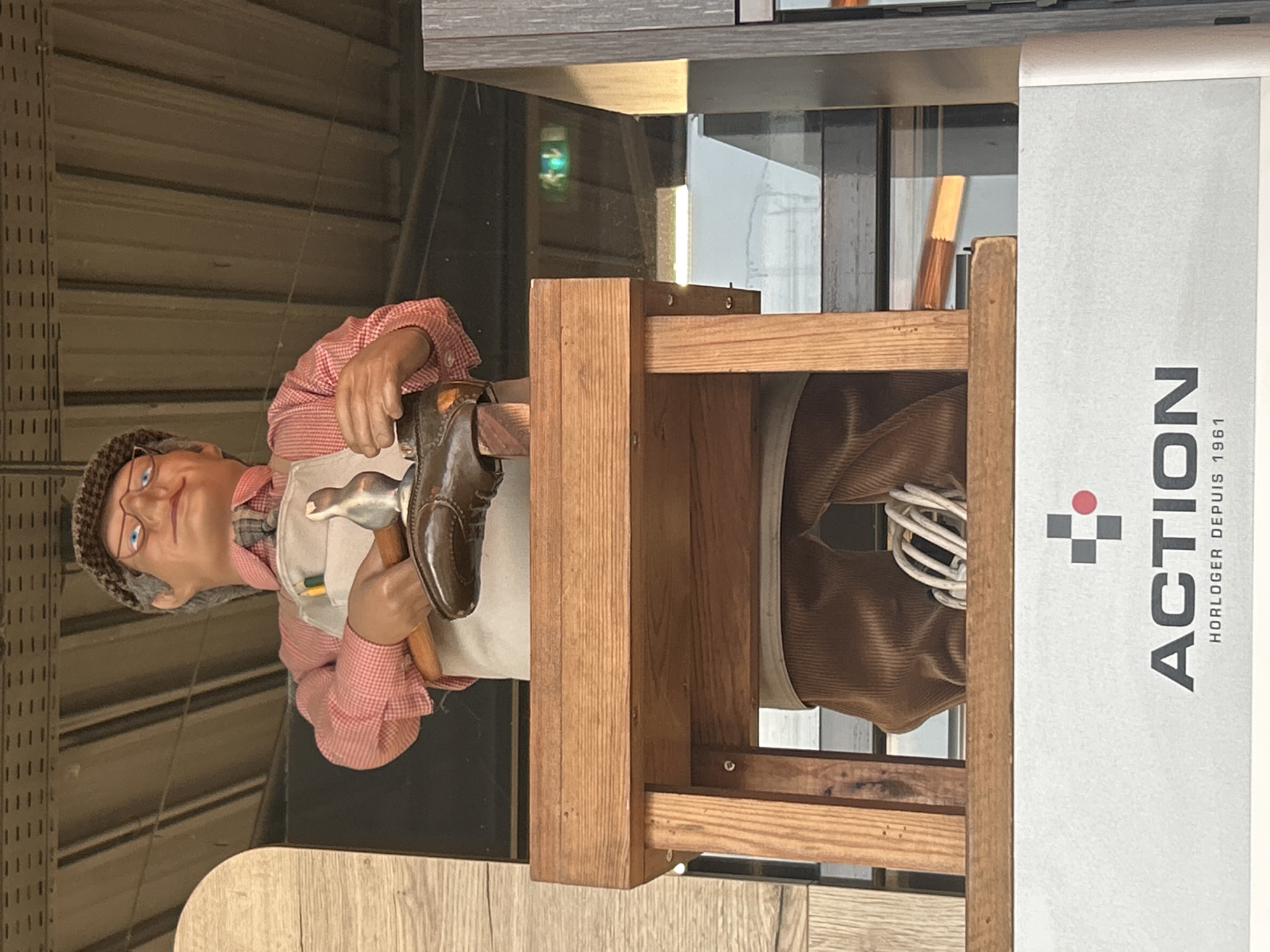 An automaton of a shoemaker stands behind a wooden workbench, holding a hammer and a brown shoe. The shoemaker wears a red and white checkered shirt, a white apron, brown corduroy pants, a flat cap, and glasses. A white cord is visible near the shoemaker's feet. Behind the automaton, a green exit sign is visible through a window. At the bottom of the image, a white sign displays the word "ACTION" in large gray letters, with smaller text below reading "HORLOGER DEPUIS 1961". The automaton and sign are likely part of a display, possibly in a shop or public space in Le Port-Marly, France.