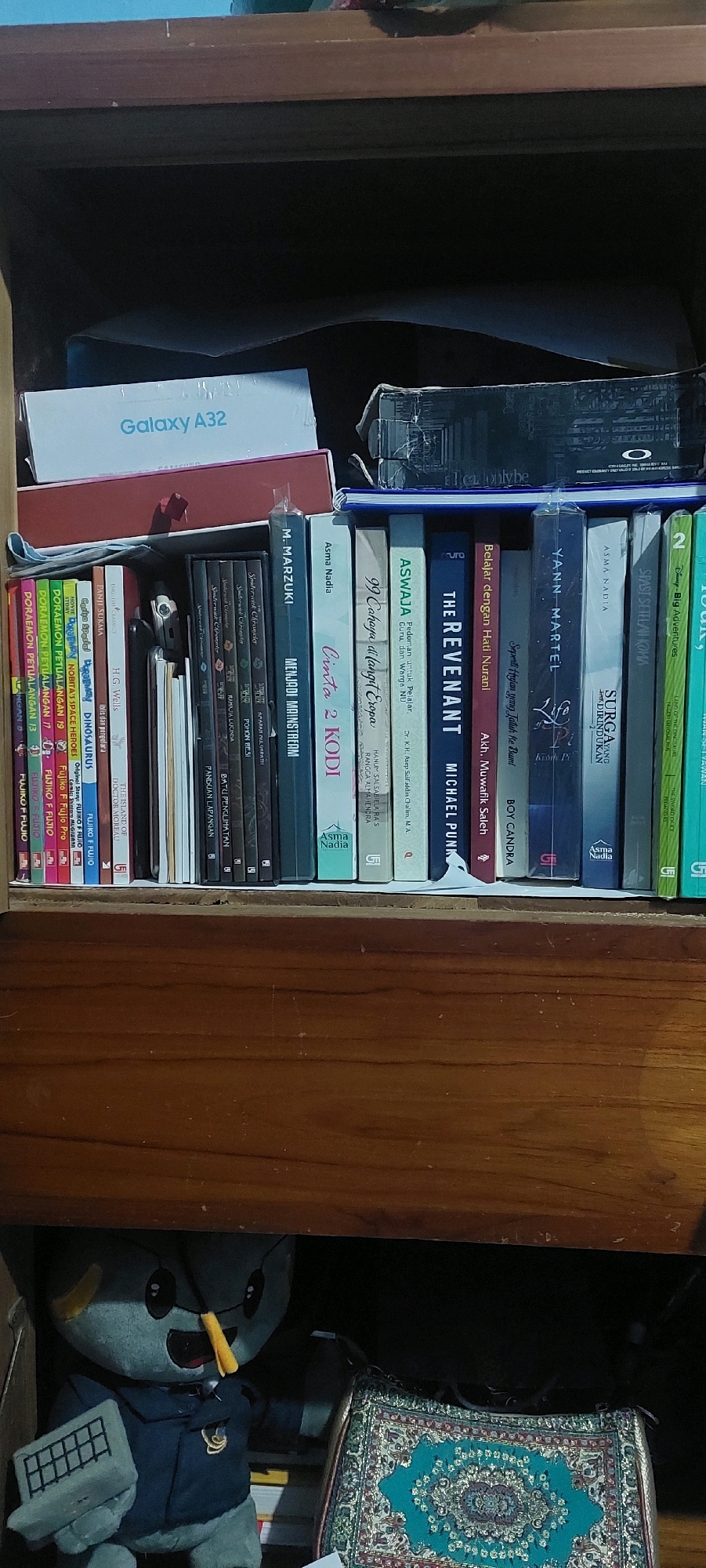 A close-up, eye-level shot shows a wooden shelf filled with books and other items. On the top shelf, a white box labeled "Galaxy A32" is visible. Below, a row of books is neatly arranged. Some book spines are in Indonesian, such as "MENJADI MAINSTREAM," "Cinta 2 KODI," "ASWAJA," and "THE REVENANT." There are also children's books with characters like "Doraemon." To the left, a grey plush toy resembling a robot with yellow accents is holding a grey object that looks like a keyboard. Next to the toy, a patterned rug with a teal background and intricate designs is partially visible. The setting appears to be an indoor shelf, possibly in a home or office, in Lumajang, Indonesia.