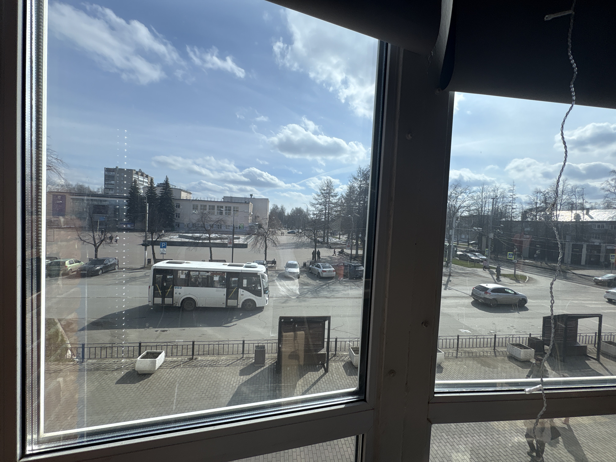View through a window of a city street. A white bus is turning at an intersection. Several cars are parked or driving on the street. Buildings and trees are visible in the background. The sky is partly cloudy. The location is Chaikovsky, Russia.