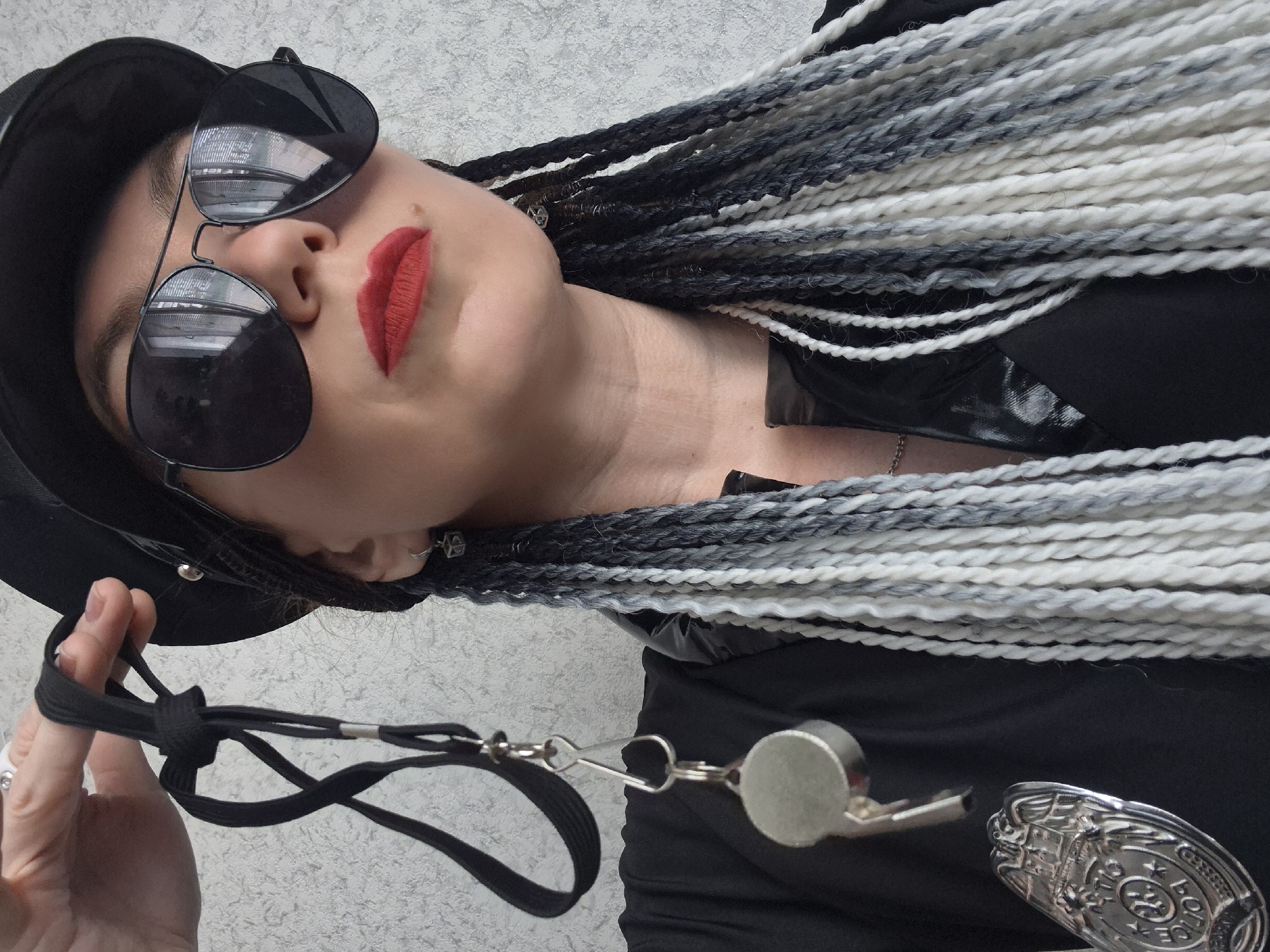 A close-up, waist-up, over-the-shoulder shot shows a woman wearing a black police-style hat, aviator sunglasses, and bright red lipstick. She has long, braided hair in shades of black, gray, and white. A lanyard with a silver whistle and a police badge hangs from her neck. The background is a textured, light-colored wall, suggesting an indoor setting in the city of Chaikovsky, Russia.