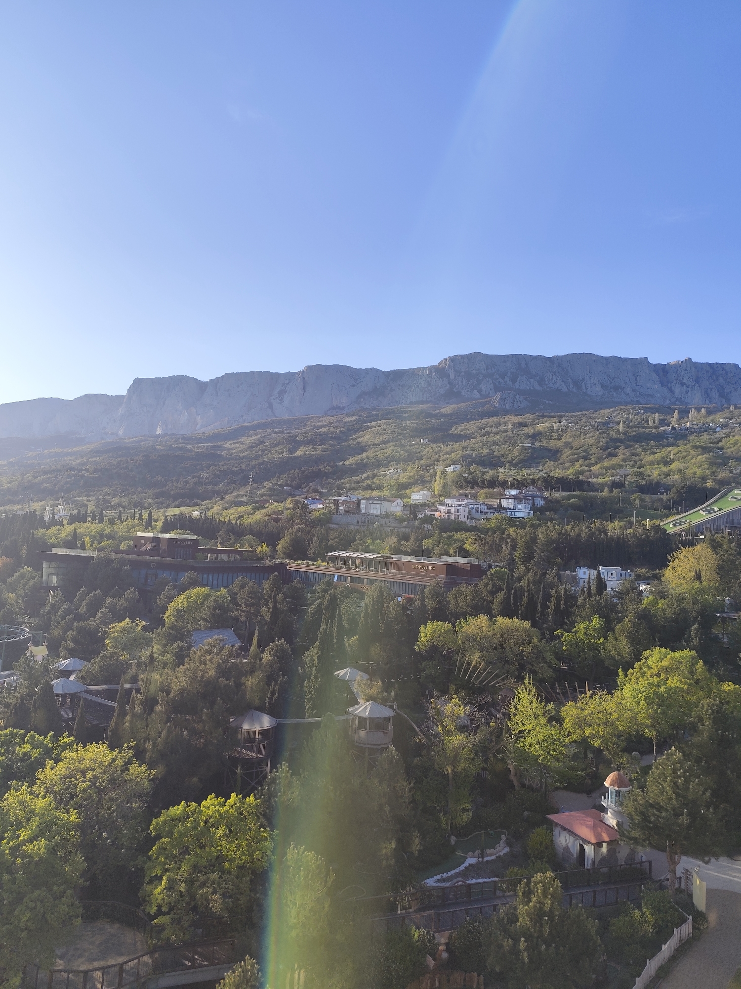 This panoramic landscape image, captured from an elevated viewpoint, presents a scenic vista dominated by mountains, lush greenery, and interspersed buildings under a clear blue sky.
In the background, a vast, rugged mountain range with gray, rocky faces extends across the horizon, partially covered by sparse vegetation. The middle ground and foreground feature a verdant hillside densely covered with a variety of trees, including tall conifers and broad-leaved deciduous trees, creating a rich green canopy.
Several buildings are nestled within this natural environment. A large, modern-looking complex with dark facades and flat roofs, possibly a resort or hotel, is prominent in the mid-ground. The text "MIRVA LIFE" is clearly visible on one section of this building. Further up the hillside, other residential-style structures with colorful roofs are scattered among the trees. In the lower right foreground, partially obscured by foliage, a building with a red tiled roof and a small, domed tower (resembling a gazebo or small lighthouse) is visible. Additionally, wooden structures that appear to be elevated walkways or treehouses are discernable amidst the foreground trees.
The sky is a bright, clear blue, indicating sunny weather. A diagonal, rainbow-colored lens flare cuts across the image, suggesting the sun is low in the sky, implying either early morning or late afternoon. No people or specific activities are visible. The scene conveys a sense of tranquility and natural beauty, characteristic of a resort or residential area within a mountainous landscape, consistent with the Симеизский поселковый совет region of Ukraine.