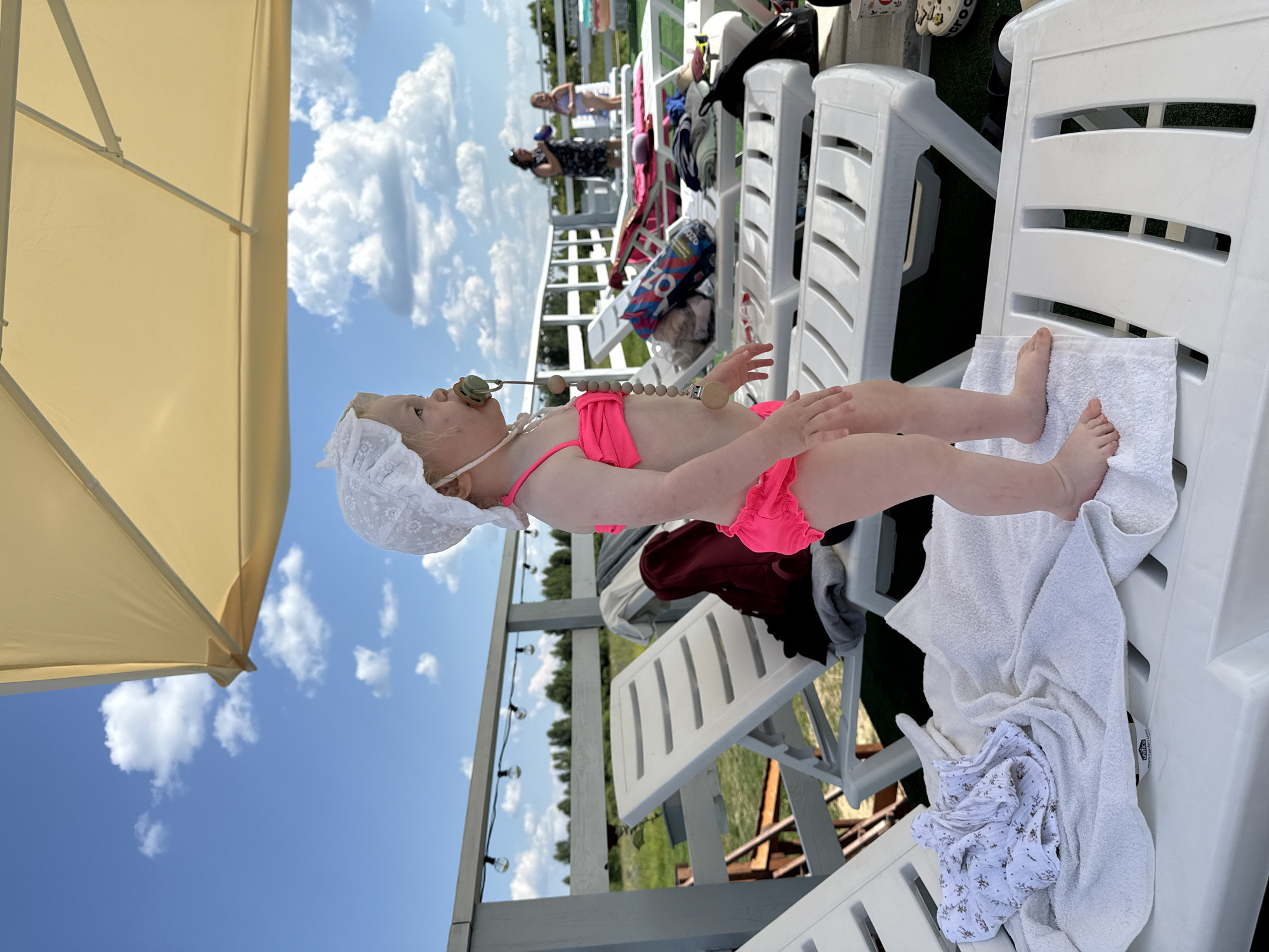 The image shows a baby lying on a white lounge chair outdoors, under a large, light-colored umbrella. The baby is wearing a white lace bonnet and a bright pink swimsuit. A pacifier is in the baby's mouth, and a beaded string with wooden beads and pacifier clips hangs around the baby's neck. The baby's legs are extended, and their feet are resting on a white towel.
In the background, several other white lounge chairs are visible, some with towels and clothing draped over them. There are also other people in the background, standing and sitting on what appears to be a deck or balcony. The scene is set against a bright blue sky with fluffy white clouds. Trees and a body of water can be seen in the distance, suggesting a waterside or park location. String lights are also visible hanging on a wooden structure.
The overall impression is a sunny, relaxed day, likely during warm weather, possibly at a resort or recreational area. The time of day appears to be daytime, with bright sunlight. No visible text is present in the image. The location context is Koloksha, Russia.