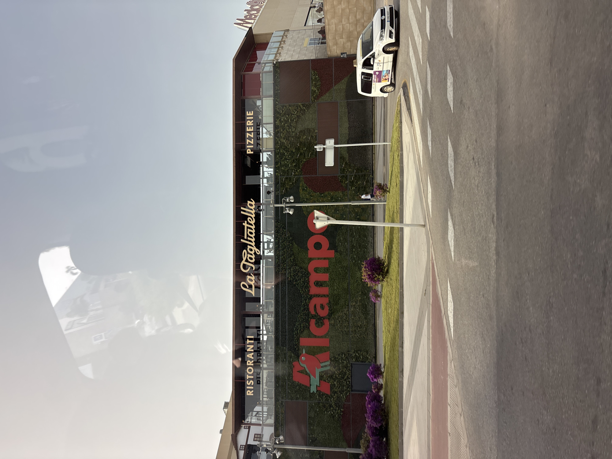 The image shows the facade of a building in Orihuela, Spain, with a large "Alcampo" logo prominently displayed against a vertical garden. Above the Alcampo logo, the building features signage for "Ristoranti," "La Tagliatella," and "Pizzerie." A white taxi is visible on the right side of the frame, stopped at a crosswalk. The sky is clear and bright, suggesting a sunny day. The building appears to be part of a commercial or shopping center, with a modern architectural style. The presence of a taxi and a crosswalk indicates a public urban setting.