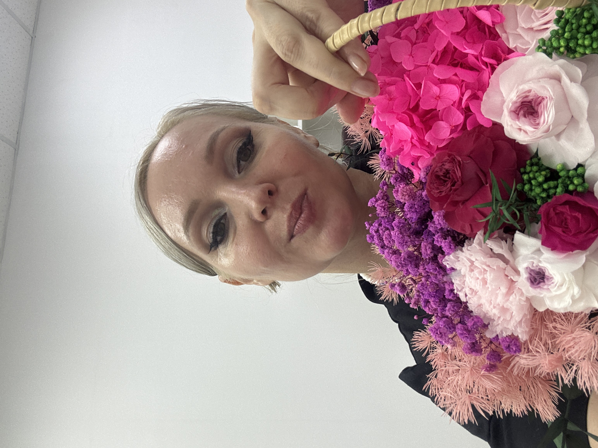 A close-up, waist-up selfie of a fair-skinned woman with blonde hair pulled back, wearing makeup and a black shirt, holding a large bouquet of vibrant pink, purple, and white flowers in a woven basket. The woman is looking at the camera with a slight smile. The background is a plain white wall with a suspended ceiling. There are no discernible geographical markers in the image to indicate a location in Komsomolsk-on-Amur, Russia.