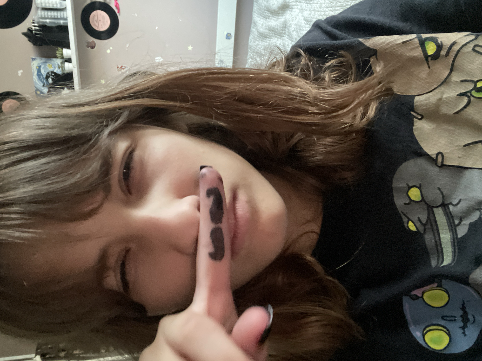 A close-up, self-taken portrait shows a young woman indoors with a fake mustache drawn on her finger. The woman has brown hair and bangs, and she is wearing a black t-shirt with cartoonish figures. Her right index finger is positioned in front of her nose, and a black mustache is drawn on the side of the finger. The background includes a shelf with items like records and small plants, suggesting a bedroom setting. There is no information to indicate the location is in Мухавецкі сельскі Савет, Belarus.