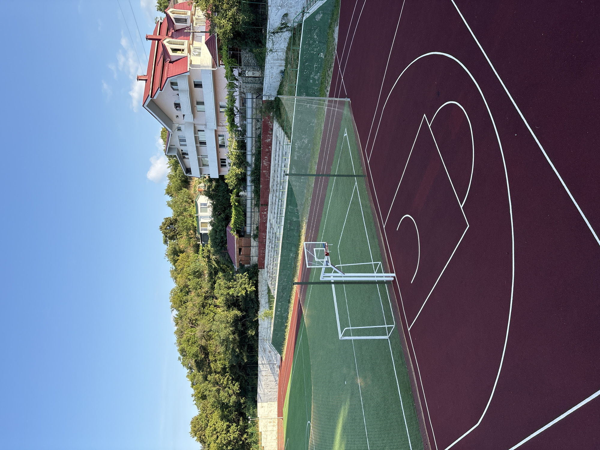 The image depicts an outdoor sports complex with a basketball court in the foreground and a soccer field with a goal behind it. The basketball court is marked with white lines on a dark red surface. The soccer field is green with white markings and a white goal. A net separates the two playing areas. In the background, there are tiered concrete bleachers and a light-colored building with a red roof, situated on a tree-covered hillside. The sky is clear and blue. The location is identified as the city of Malyi Mayak, Ukraine. There are no people visible in the image.