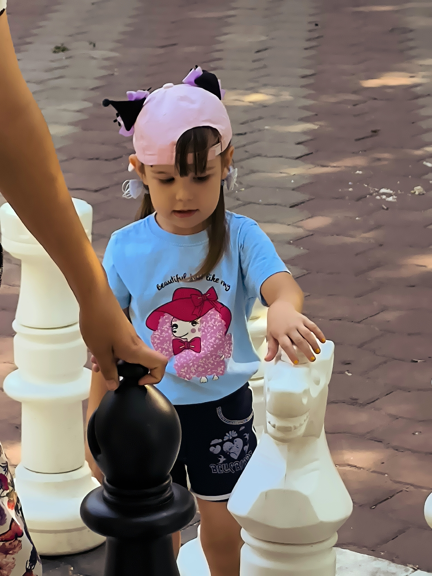 In Karaganda, Kazakhstan, a young girl wearing a pink baseball cap with cat ears and a blue t-shirt with a poodle graphic is engaged in a game of giant chess. Her hand, with bright yellow nail polish, rests on a white chess piece shaped like a horse. Another person's arm, holding a black chess piece, is visible on the left side of the frame, suggesting they are playing with the girl. The game is taking place outdoors on a paved area, possibly a park or plaza, with textured brown paving stones.