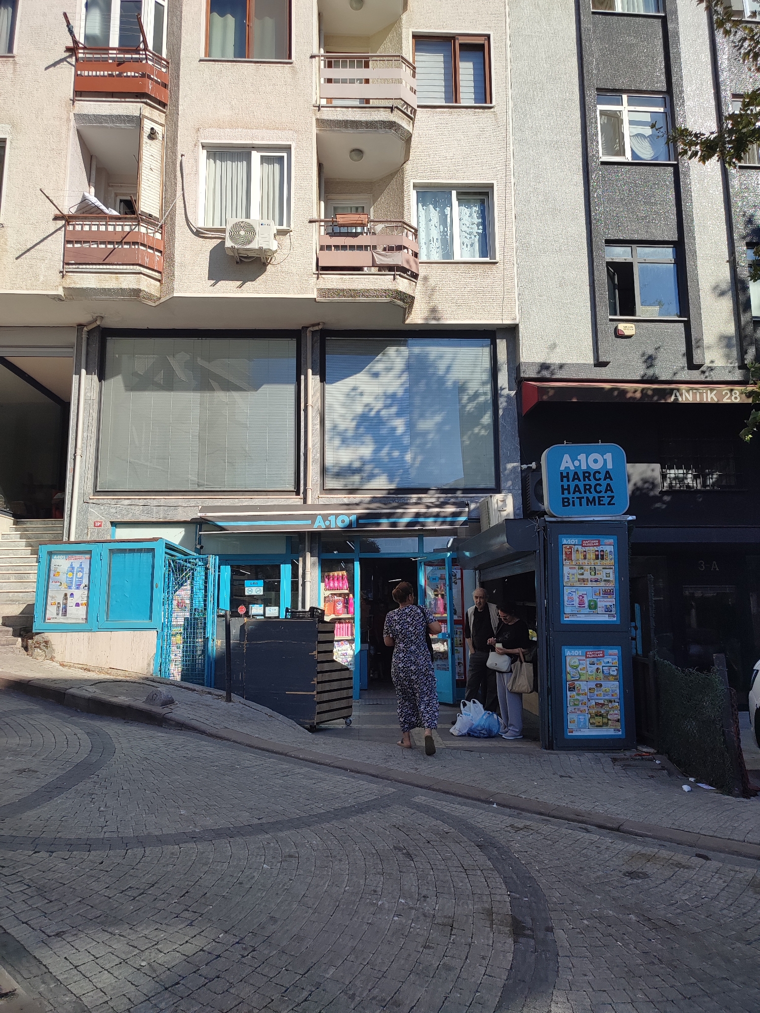 The image depicts a street scene in Üsküdar, Turkey, outside an A101 store. In the foreground, a cobblestone street curves upwards to the right. A blue kiosk-like structure stands on the left side of the street, displaying product advertisements. Adjacent to it is the entrance to the A101 store, with a blue awning and large glass windows. A woman in a patterned dress is walking away from the store towards the viewer. Two other individuals, a man and a woman, are standing near the store entrance, possibly entering or exiting. Behind the kiosk and store, a multi-story building is visible, with balconies and windows. The building to the right of the A101 store has a darker facade with the text "ANTIK 28" visible. The scene appears to be during daylight, with shadows cast on the street and buildings.