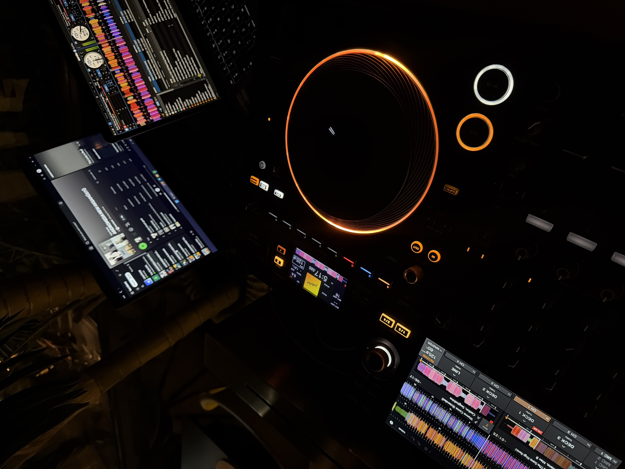 The image displays a dimly lit DJ setup, likely a performance or practice space, set at night in Budva, Montenegro. The central subject is a professional DJ controller, prominently featuring an illuminated orange jog wheel, suggesting an active session. Several bright buttons are visible, including "CUE," "SLIP REV," "IN," and "OUT," along with various knobs and faders.
A small display screen on the controller shows track information: "5:17.525" duration, "125.9" BPM, and the track "Wanna Come (Extended)" by "Sergiodnine."
Above the controller, a laptop screen displays "Serato DJ Pro" software, characterized by vivid, multi-colored waveforms in shades of pink, red, orange, yellow, green, blue, and purple. These waveforms represent tracks assigned to "DECK 1" and "DECK 2." Visible track titles include "Wanna Come (Extended)" by "Castion, Sergiodnine" and a remix of a track by "tana, Max B." The screen also shows BPMs around "125.9," time remaining/elapsed (e.g., "-05:17," "-01:25"), and deck status, including an "ON AIR" indicator for "DECK 1."
To the left, a second screen, possibly a tablet or another laptop, displays what appears to be a music library or streaming service interface, showing text like "ANDROMEDA NEW AKSAMÜSTÜ" and "ANDROMEDANER," alongside timestamps such as "22:18:03 CEST."
The scene emphasizes the technological aspect of DJing, with no people visible. The bright, colorful lights emanating from the screens and the controller contrast sharply with the dark surroundings, creating an immersive atmosphere typical of a DJ booth or studio environment.