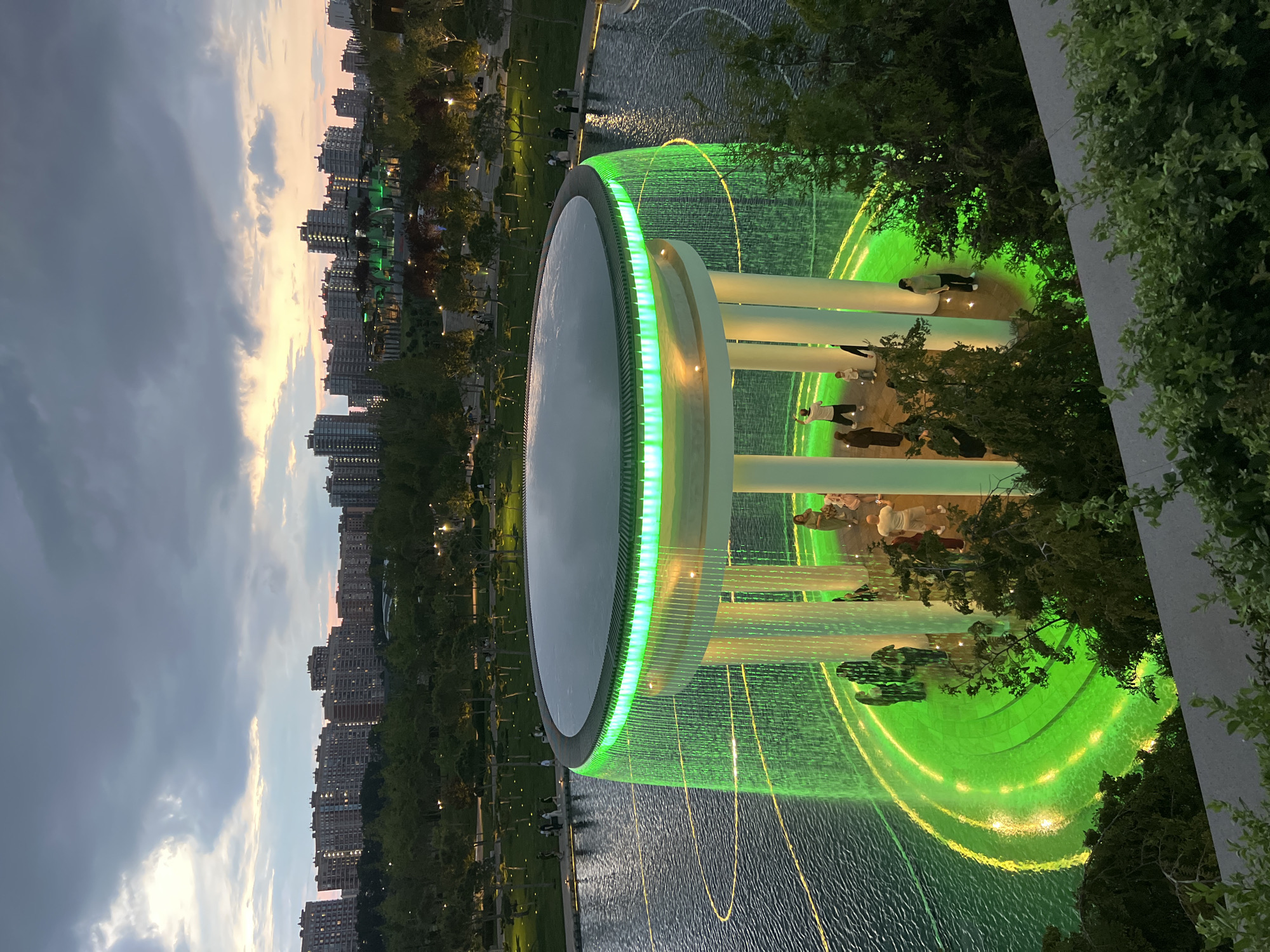 The image depicts a nighttime scene at the Fountain of Krasnodar (also known as the "Flower of Life" fountain) located in Krasnodar, Russia. A circular structure with white columns is illuminated by green lights, and water cascades down from its rim, creating a curtain-like effect. The water falls into a pool with concentric rings of green light. Several people are gathered around and under the structure, some observing the fountain. In the background, a cityscape with tall buildings is visible against a dusky sky, with lights on in many of the structures. The surrounding area appears to be a park with trees and pathways, also lit by artificial lights. The overall atmosphere is one of a modern urban public space at dusk.
