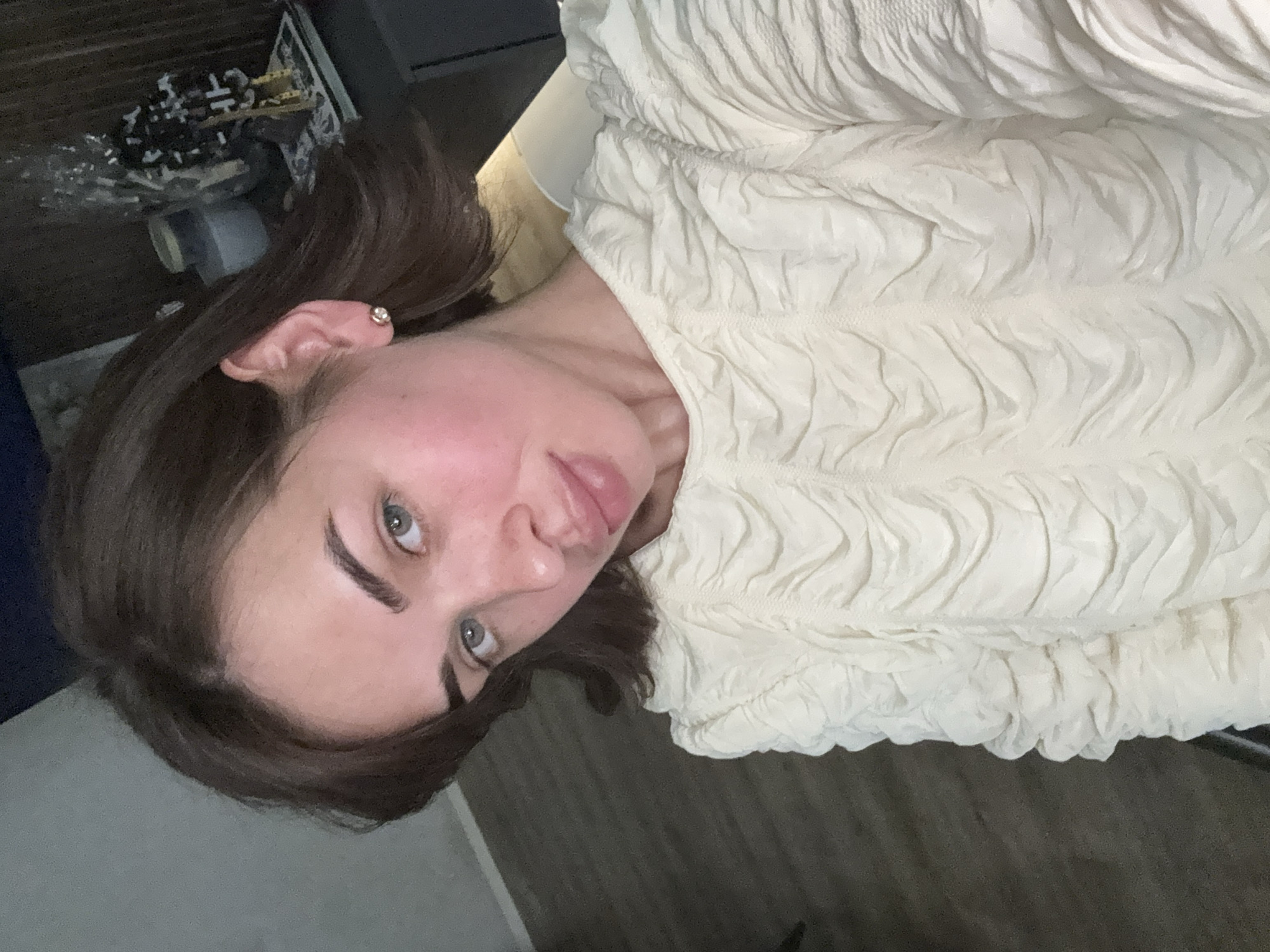 A woman with shoulder-length brown hair is wearing a textured off-white top. She has fair skin, green eyes, and prominent eyebrows. She is wearing a small, round earring in her left ear. The background includes a glimpse of a desk with items on it, and what appears to be a dark floor. The location is not discernible as Лобаново, Russia based solely on the visible elements in the image. There is no verifiable information about the location available in the image.