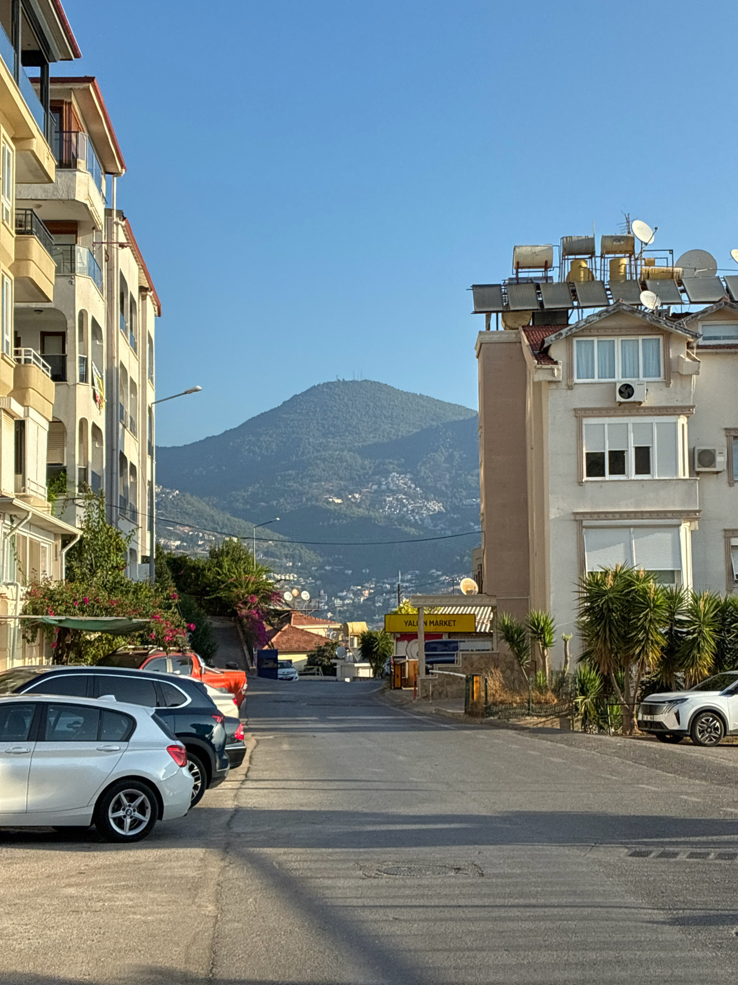 The image shows a street scene in Alanya, Turkey. The street is lined with buildings on both sides, with cars parked along the side of the road. In the background, a large, tree-covered mountain dominates the landscape. The sky is clear and blue, indicating a sunny day. The buildings appear to be apartment buildings or hotels, with balconies and windows visible. Solar panels and satellite dishes are mounted on the roofs of some of the buildings. The street itself is paved and appears to be in good condition. A yellow sign with the text "YALIN MARKET" is visible on the right side of the street. The overall impression is one of a warm, sunny day in a Mediterranean coastal city.