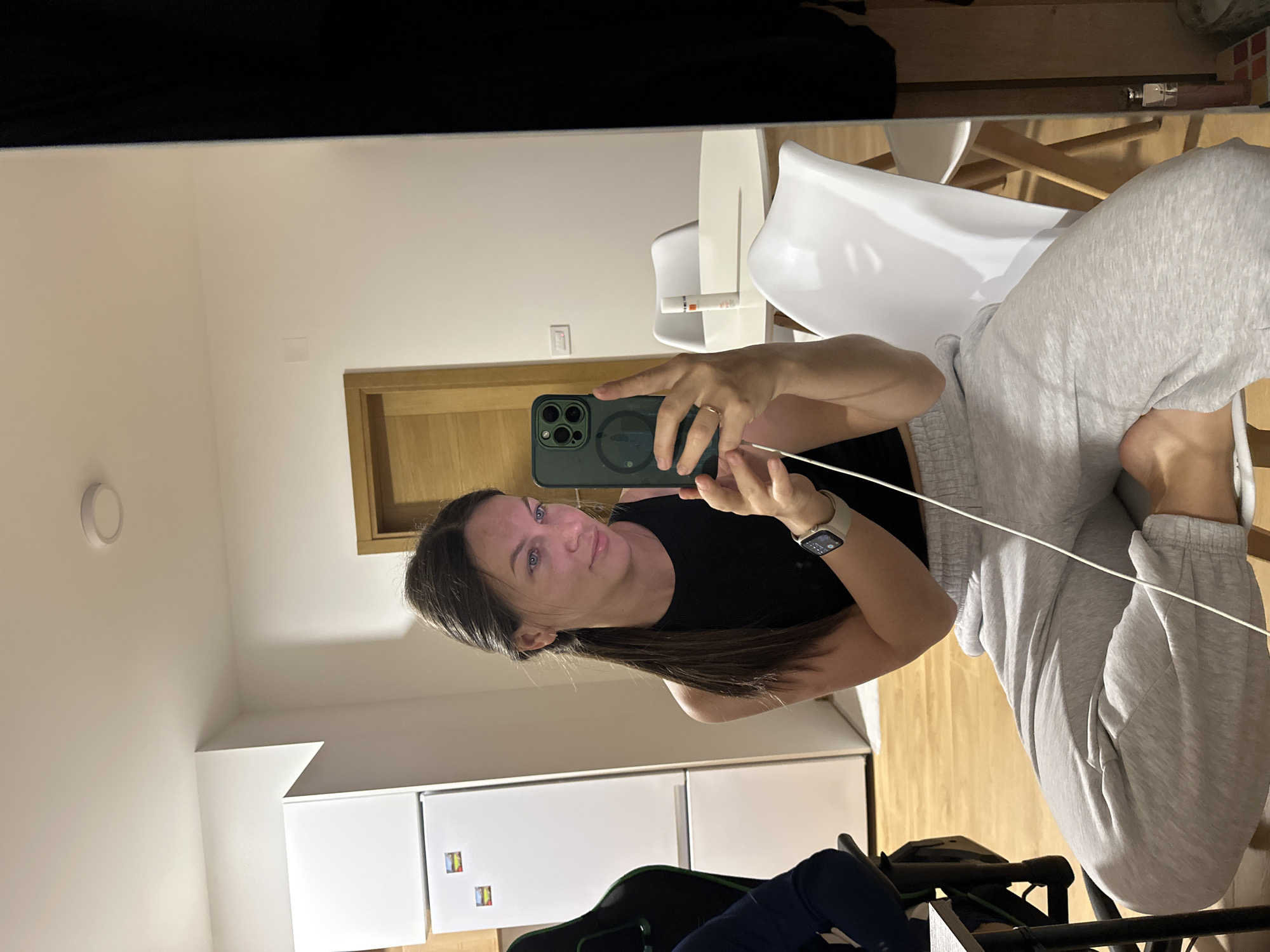 A woman is taking a selfie indoors. She's wearing a black tank top and gray sweatpants, and an Apple watch on her left wrist. She's holding a dark green iPhone with a circular camera array. In the background, there's a white wall, a wooden door frame, a white refrigerator, and a white high chair. There is also a gaming chair and wooden floor. The location is Donji Pobori, Montenegro.